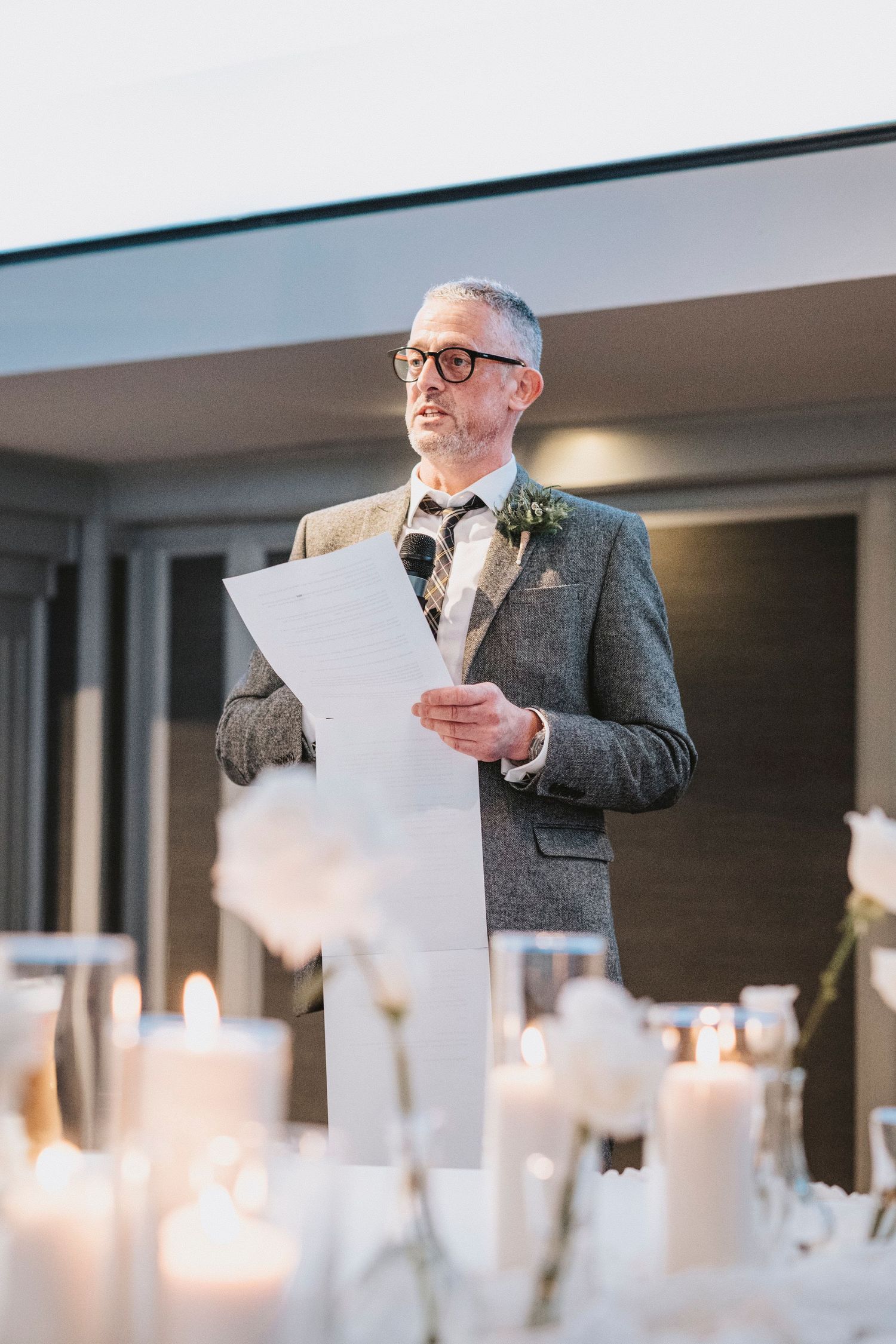 A person in glasses and grey suit gives a speech at an elegant candlelit event while reading from papers.