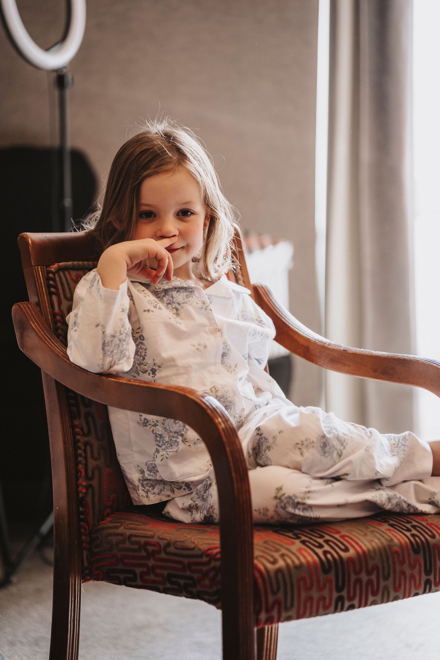 A child in a white lace dress lounges playfully across a vintage wooden armchair with geometric upholstery.