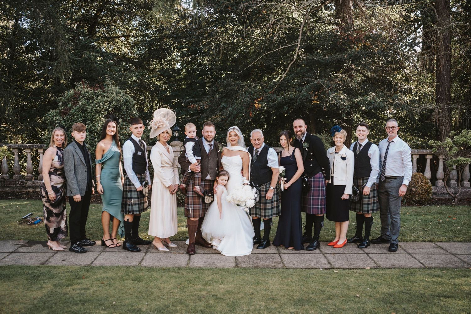Wedding party members stand in a line outdoors against a wooded backdrop for group photos.