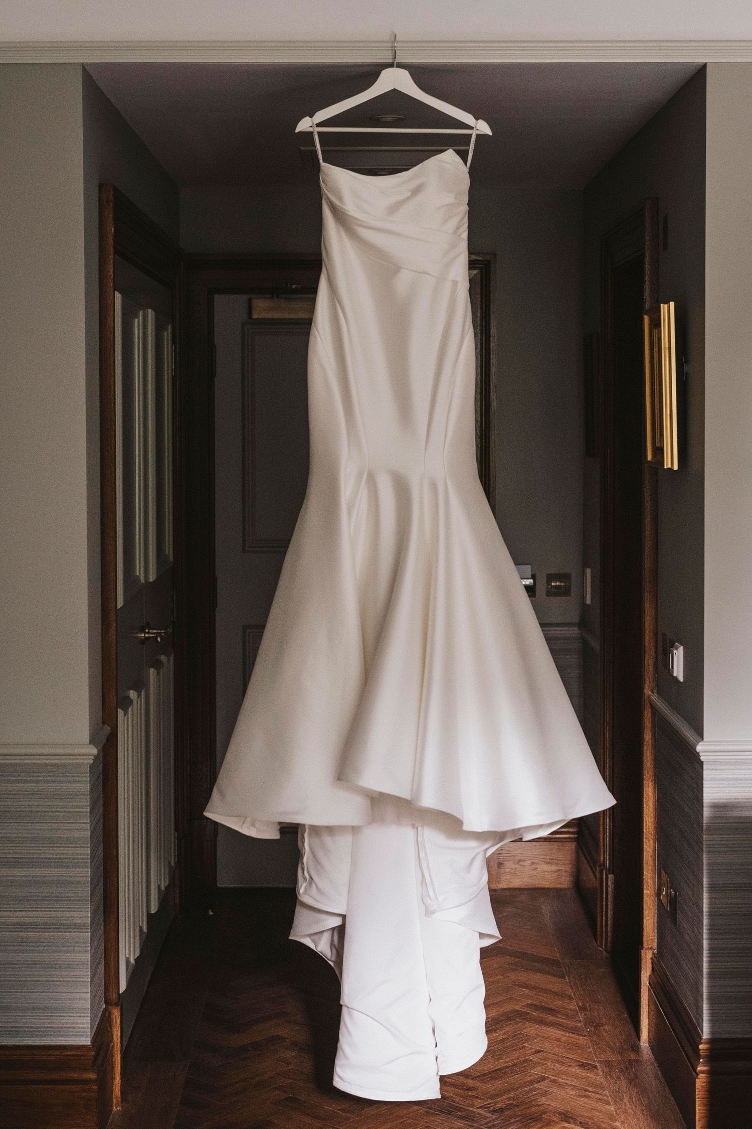 An elegant white mermaid-style wedding dress hangs on display in a dark hallway with hardwood flooring.