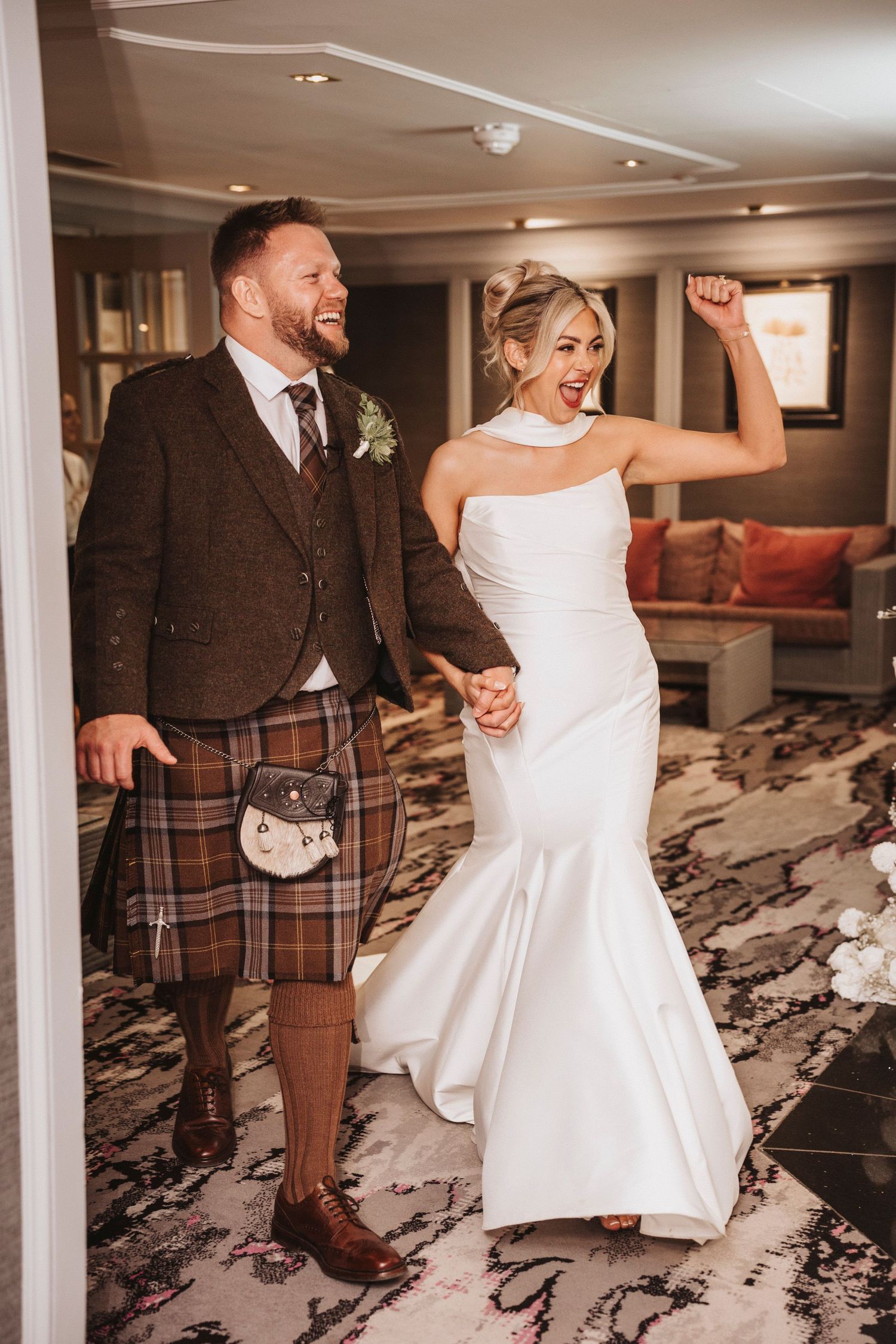 A couple in wedding attire dances joyfully in an elegant room with patterned carpeting.