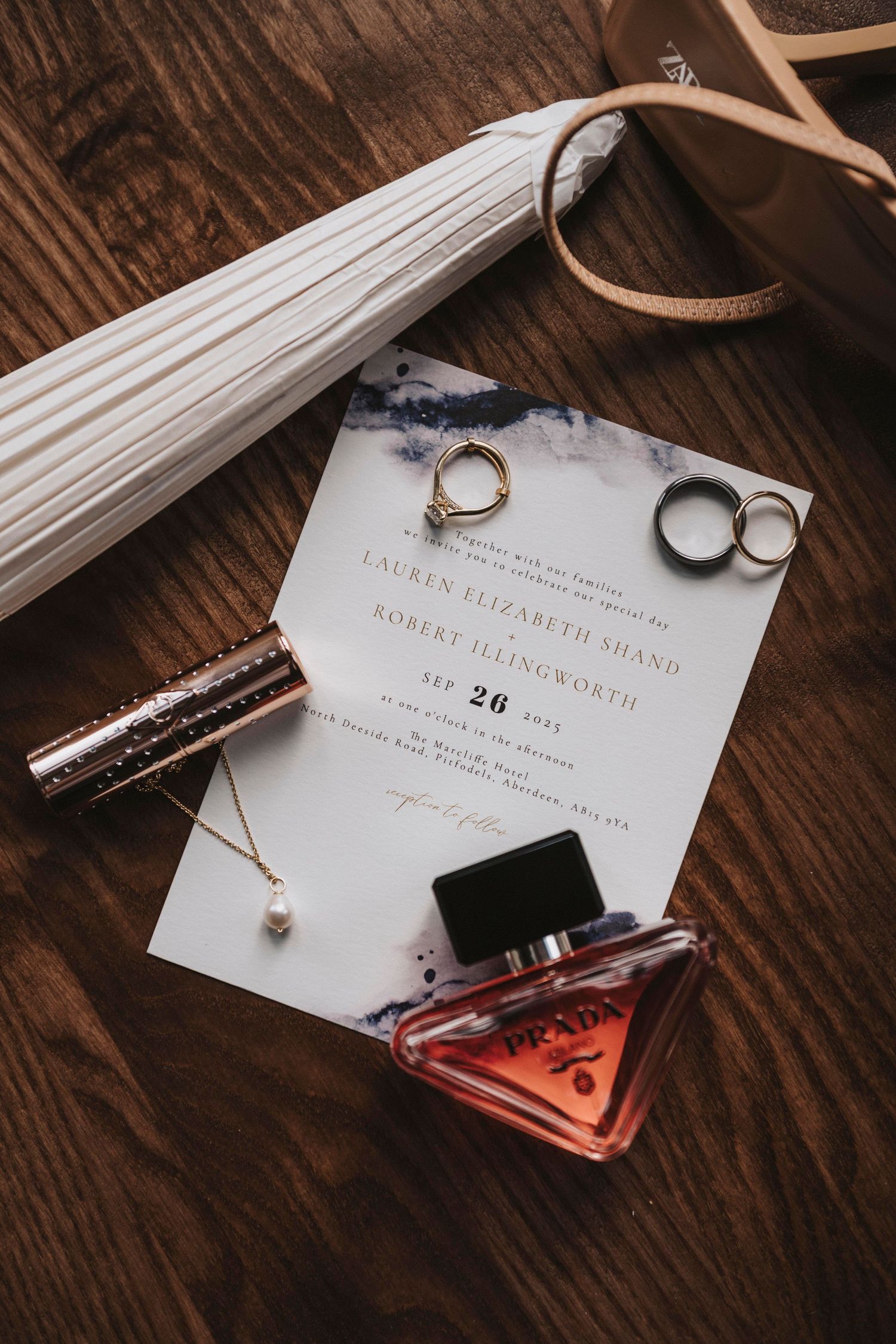 Wedding details including invitation, rings, perfume bottle, and tassel on rustic wooden surface in warm lighting.