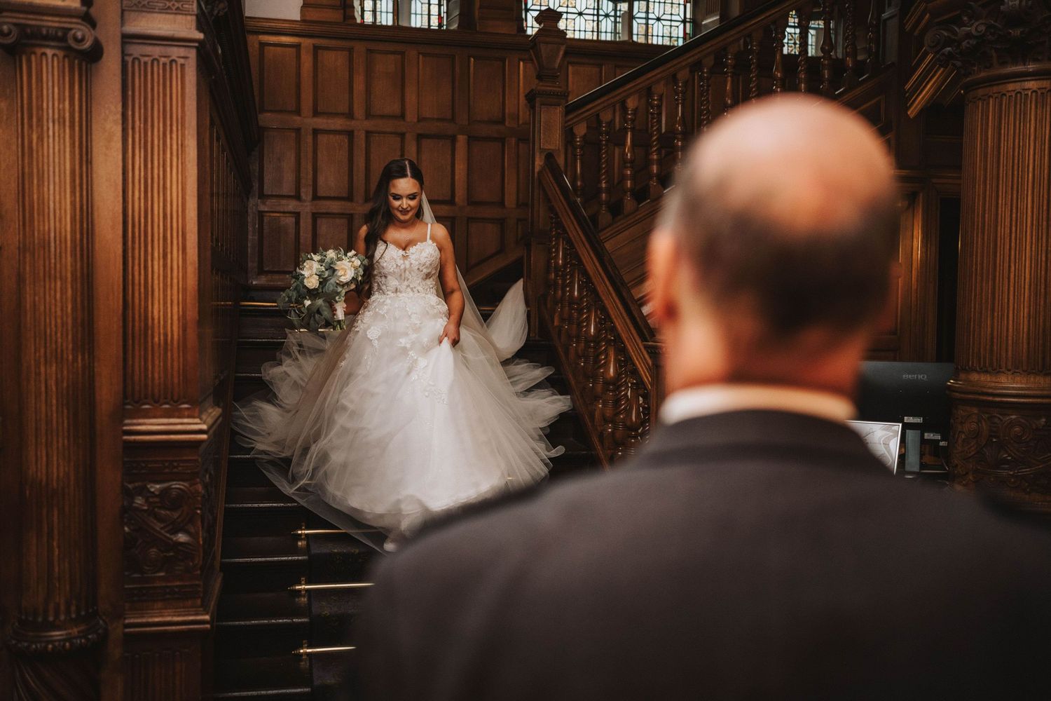 A couple walks up a grand wooden staircase in a dark-toned historic building with ornate architectural details.