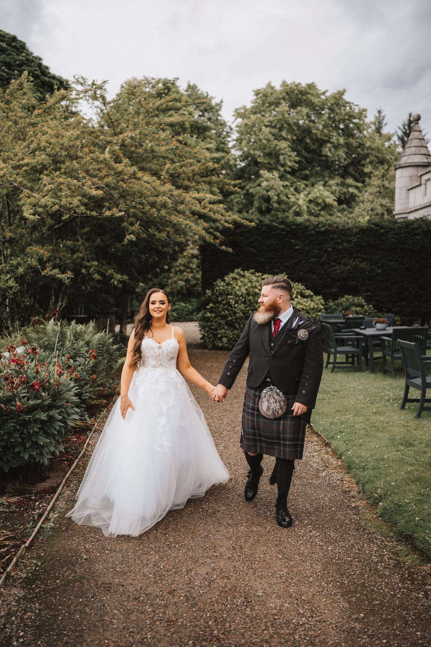 A couple walks along a tree-lined garden path with the bride wearing a flowing white gown.