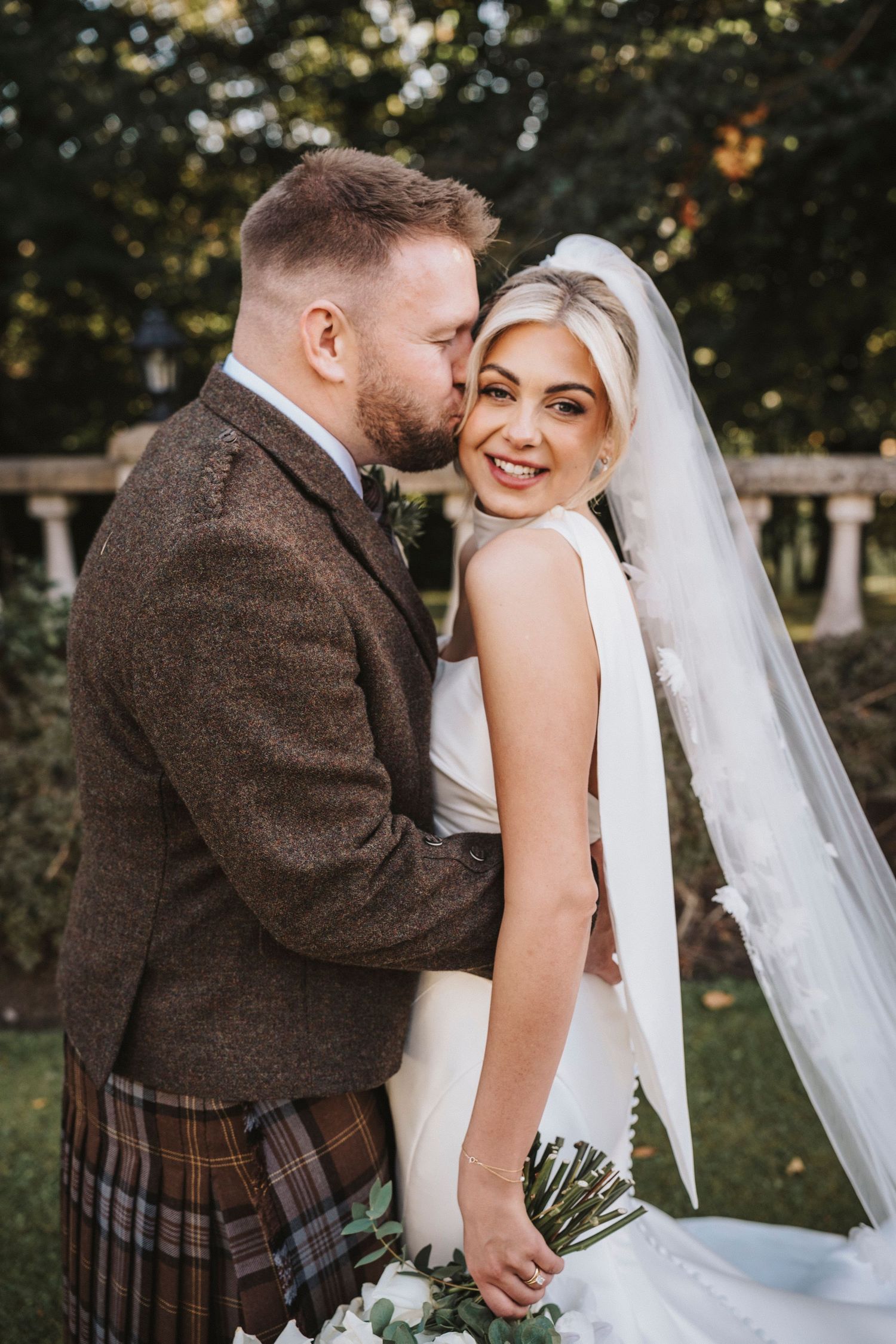 Romantic wedding couple embracing outdoors with bride in white dress and groom in traditional Scottish attire.