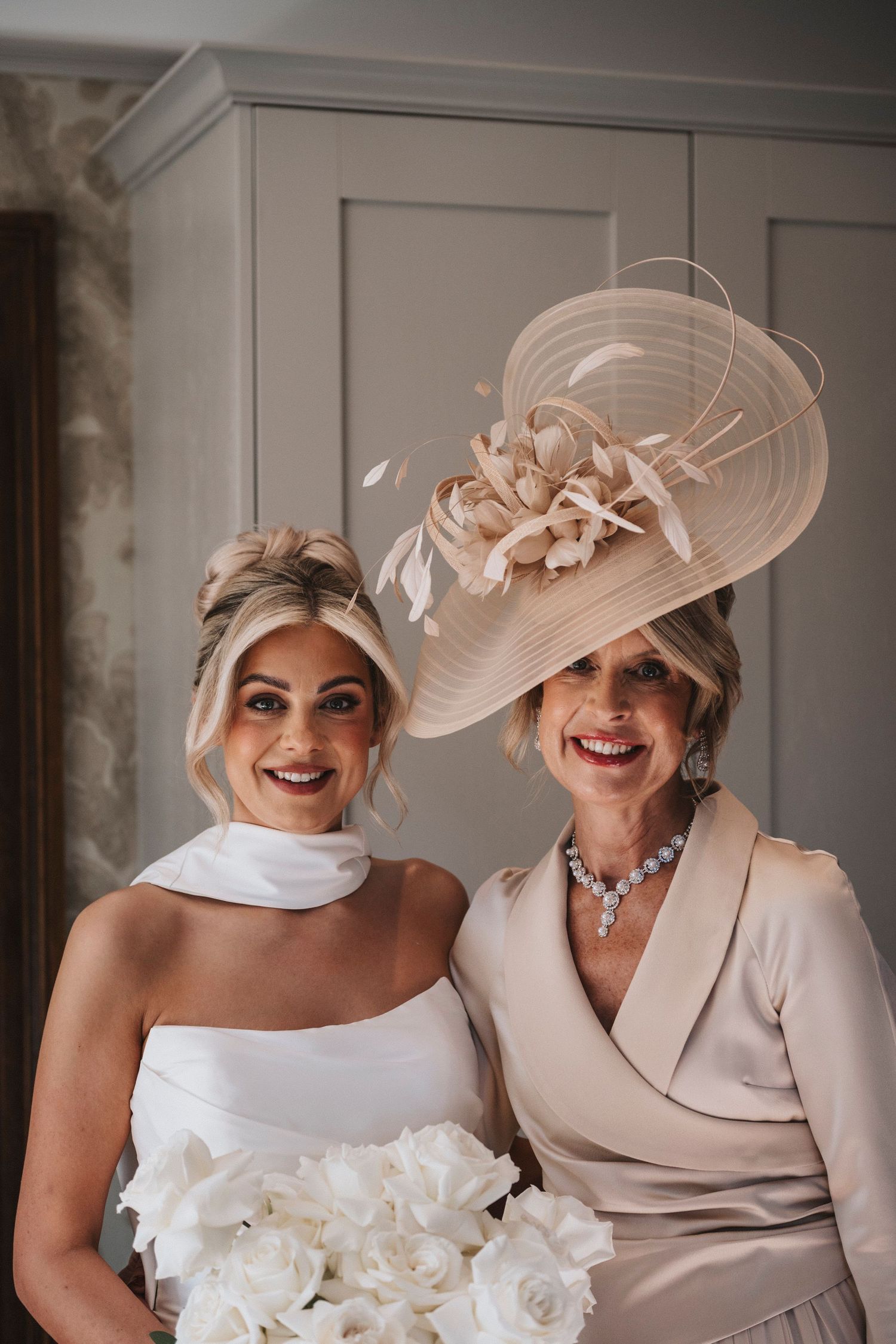 Two elegant women in formal attire wearing matching blush-colored wide-brimmed hats share joyful laughter at a special occasion.