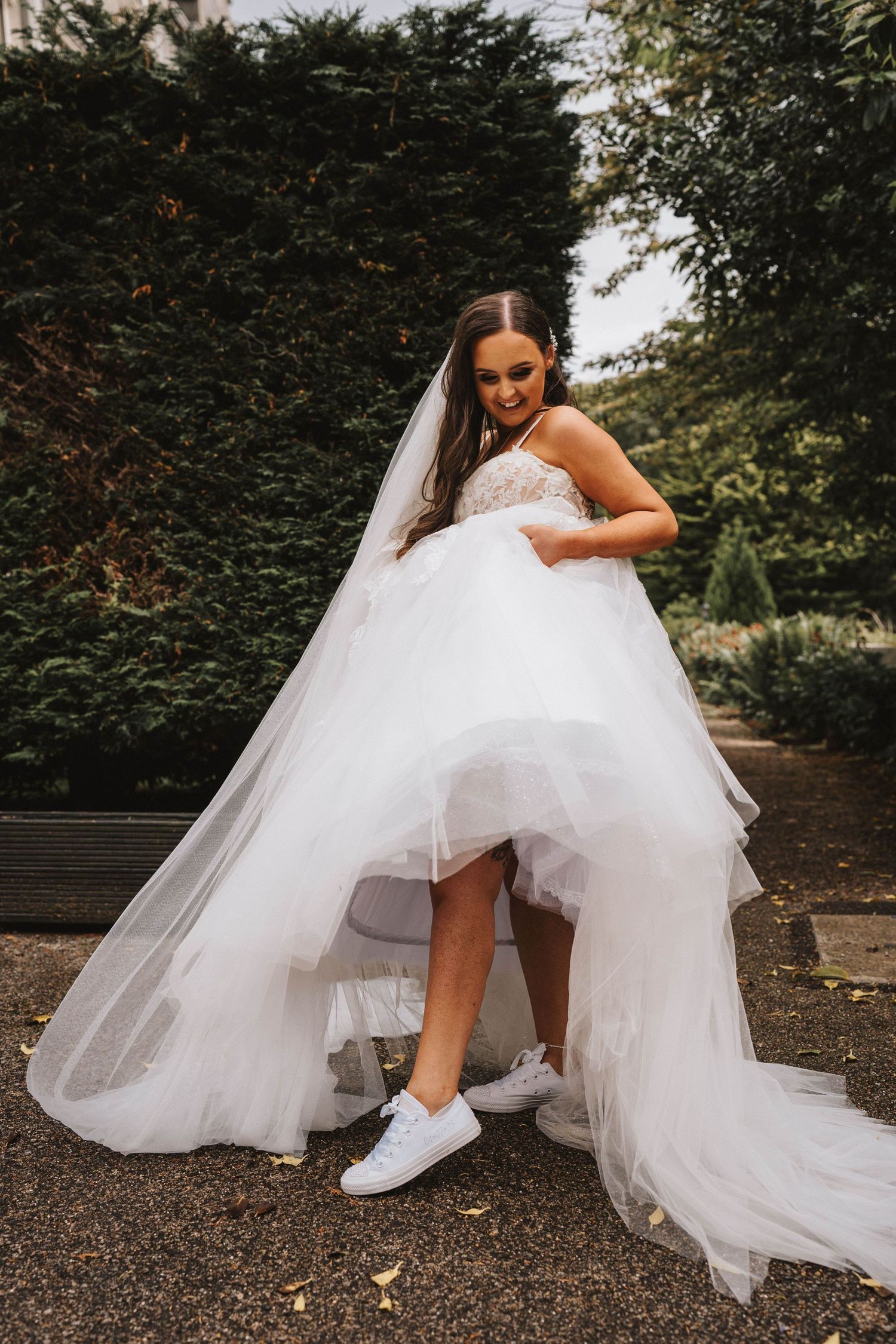 Playful bride in white wedding dress and sneakers showing off her outfit in a garden setting.