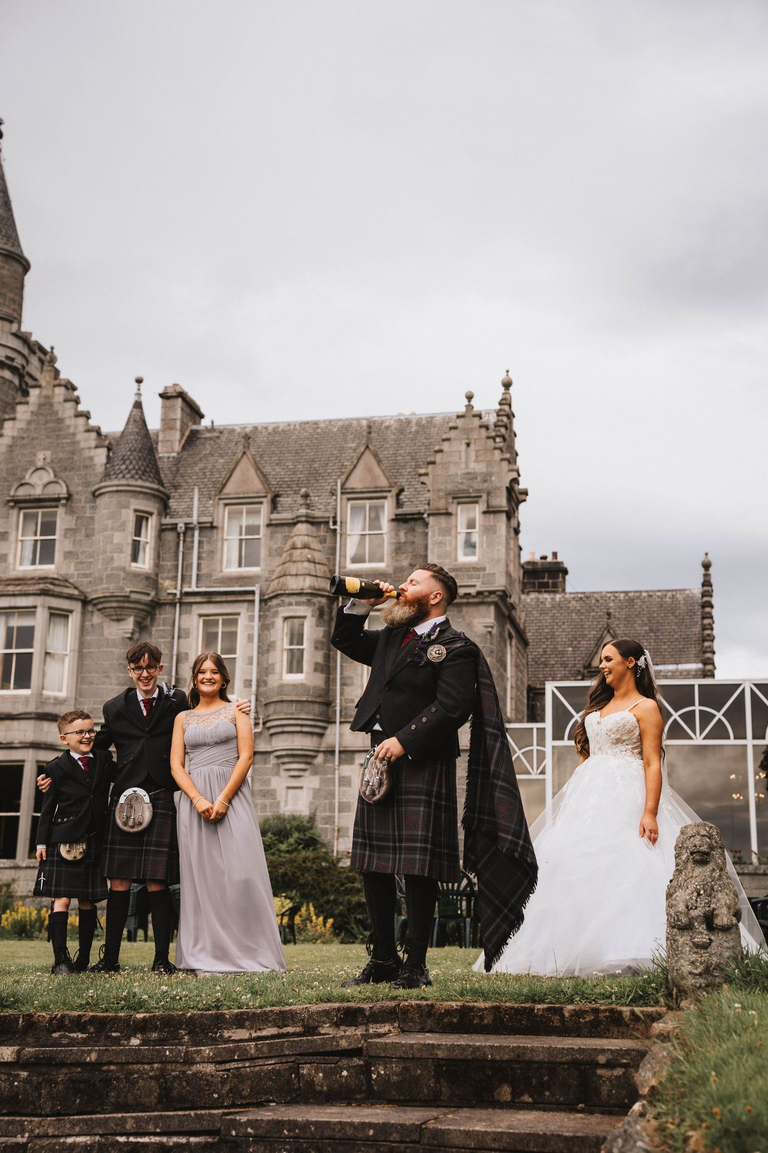 Wedding party gathers for photos outside historic Scottish stone mansion.