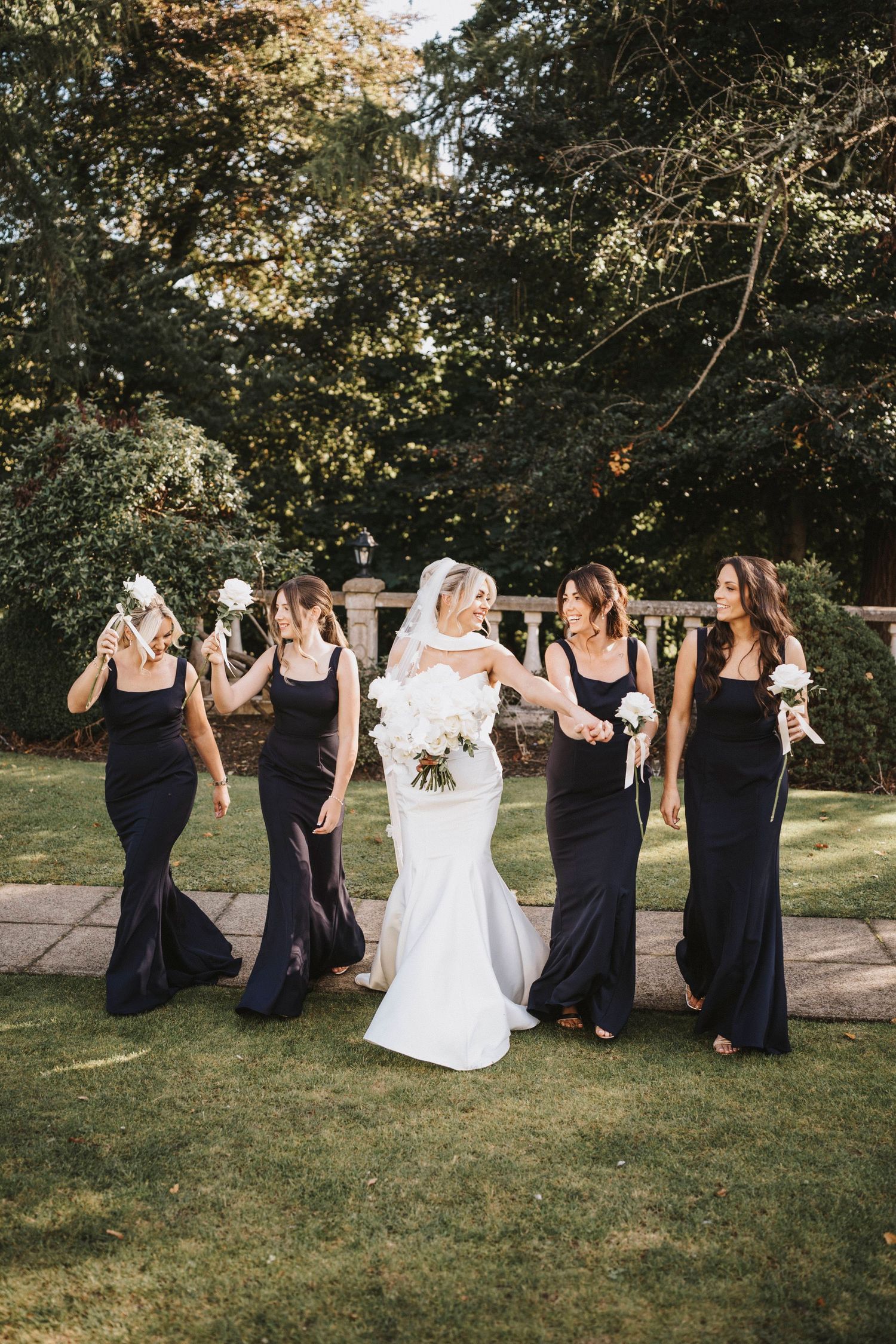 A bridal party in black dresses and white gown walk together in a line on grassy lawn during outdoor wedding celebration.