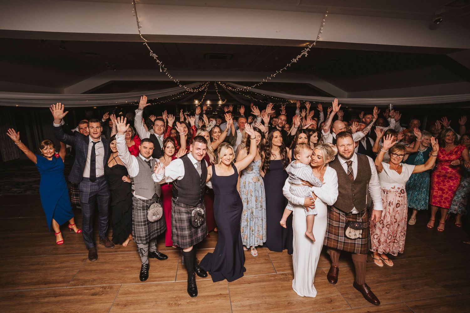 Large group of wedding guests dancing with their arms raised on a wooden dance floor at a reception celebration.