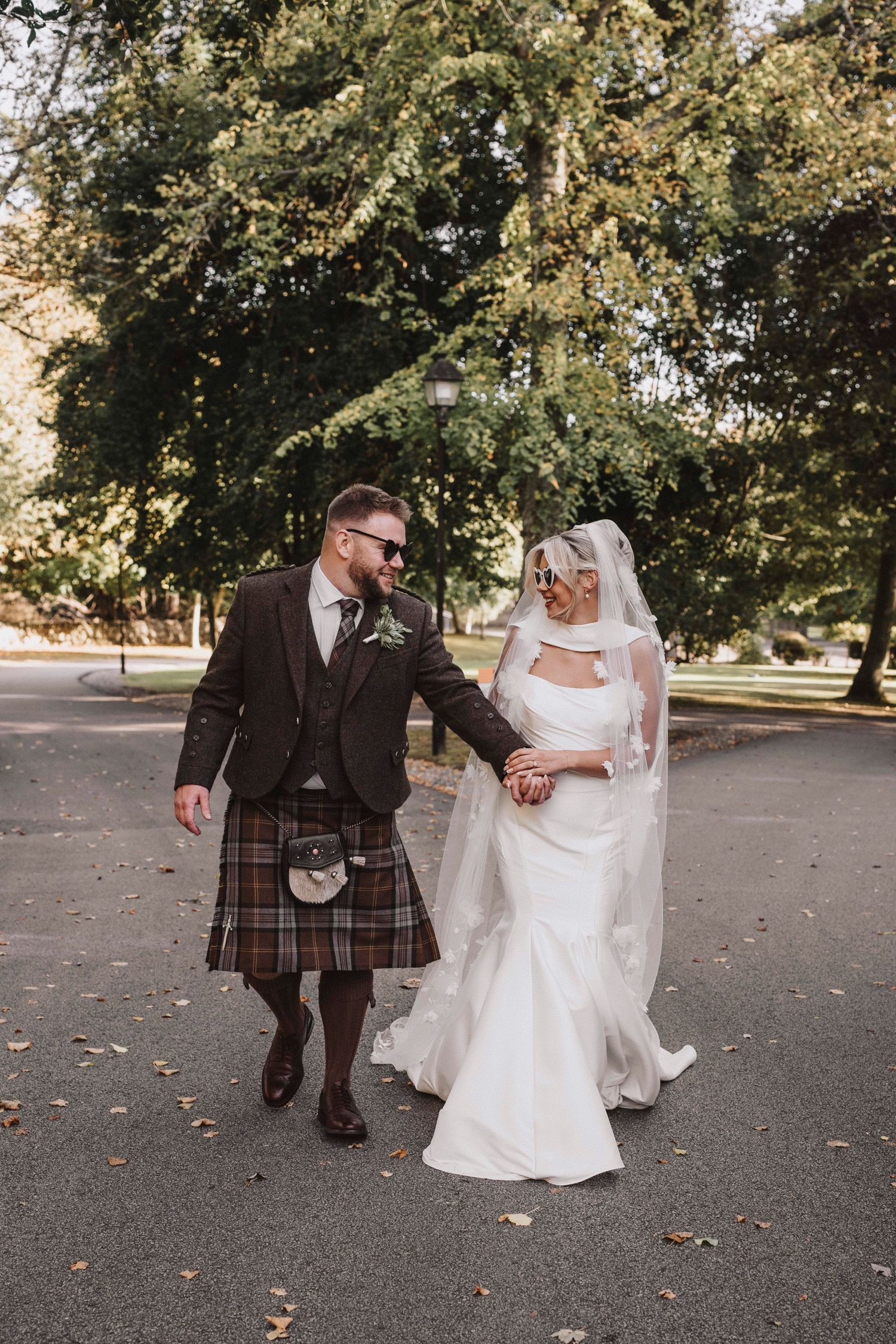 A couple in wedding attire walk together on a path covered in autumn leaves, with the groom wearing a traditional kilt.