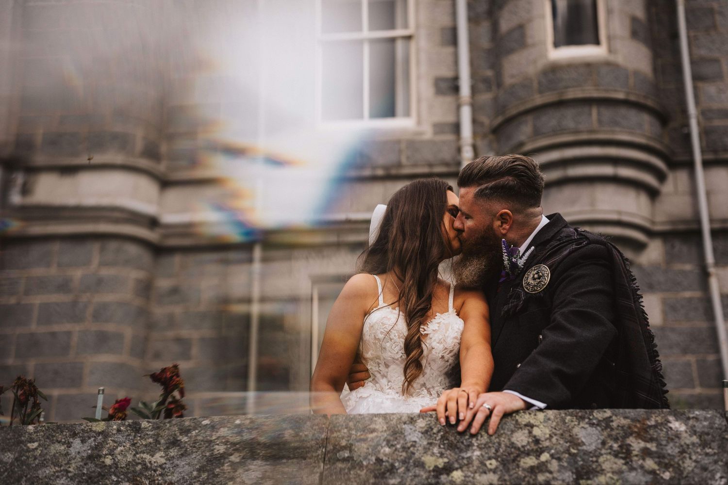 A couple in wedding attire shares an intimate moment on stone steps outside a historic stone building.