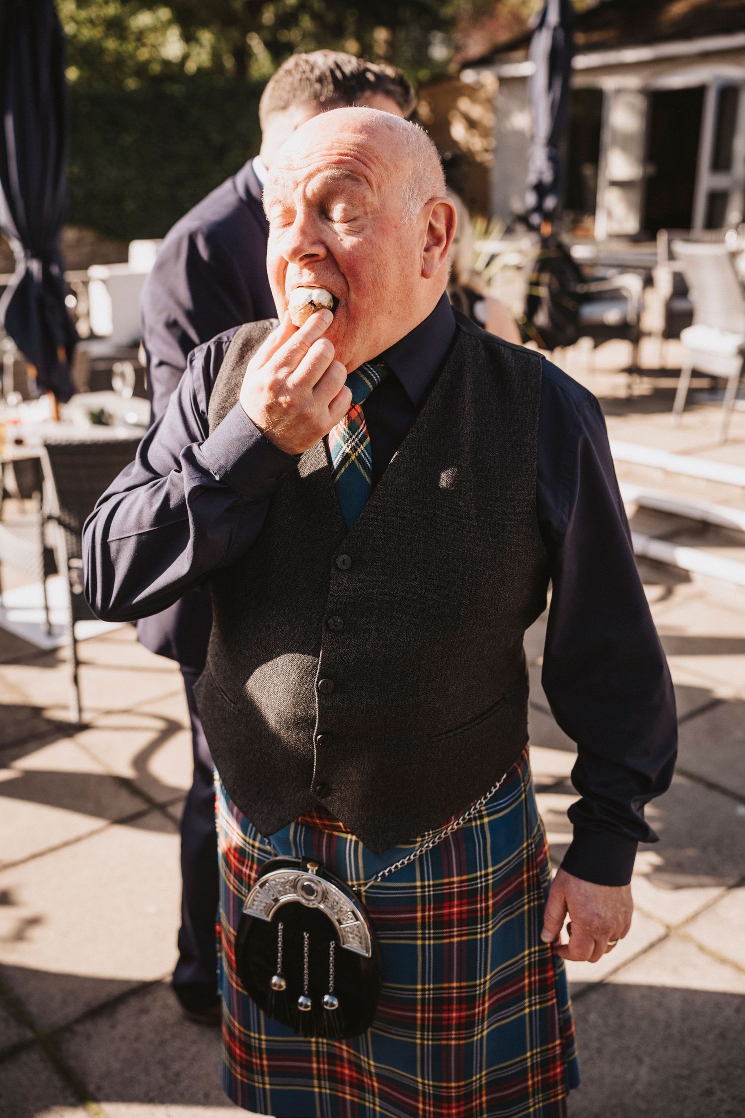 A person wearing a traditional black sweater and tartan kilt laughs while standing outdoors in sunlight.