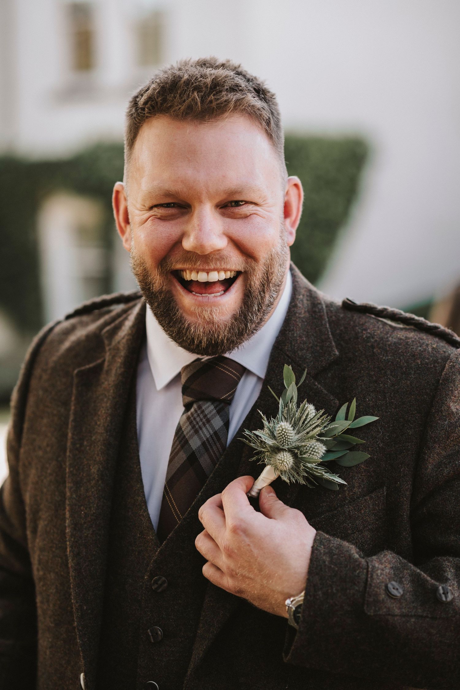 A groom in a dark suit and tie laughs joyfully while wearing a succulent boutonniere at an outdoor wedding venue.