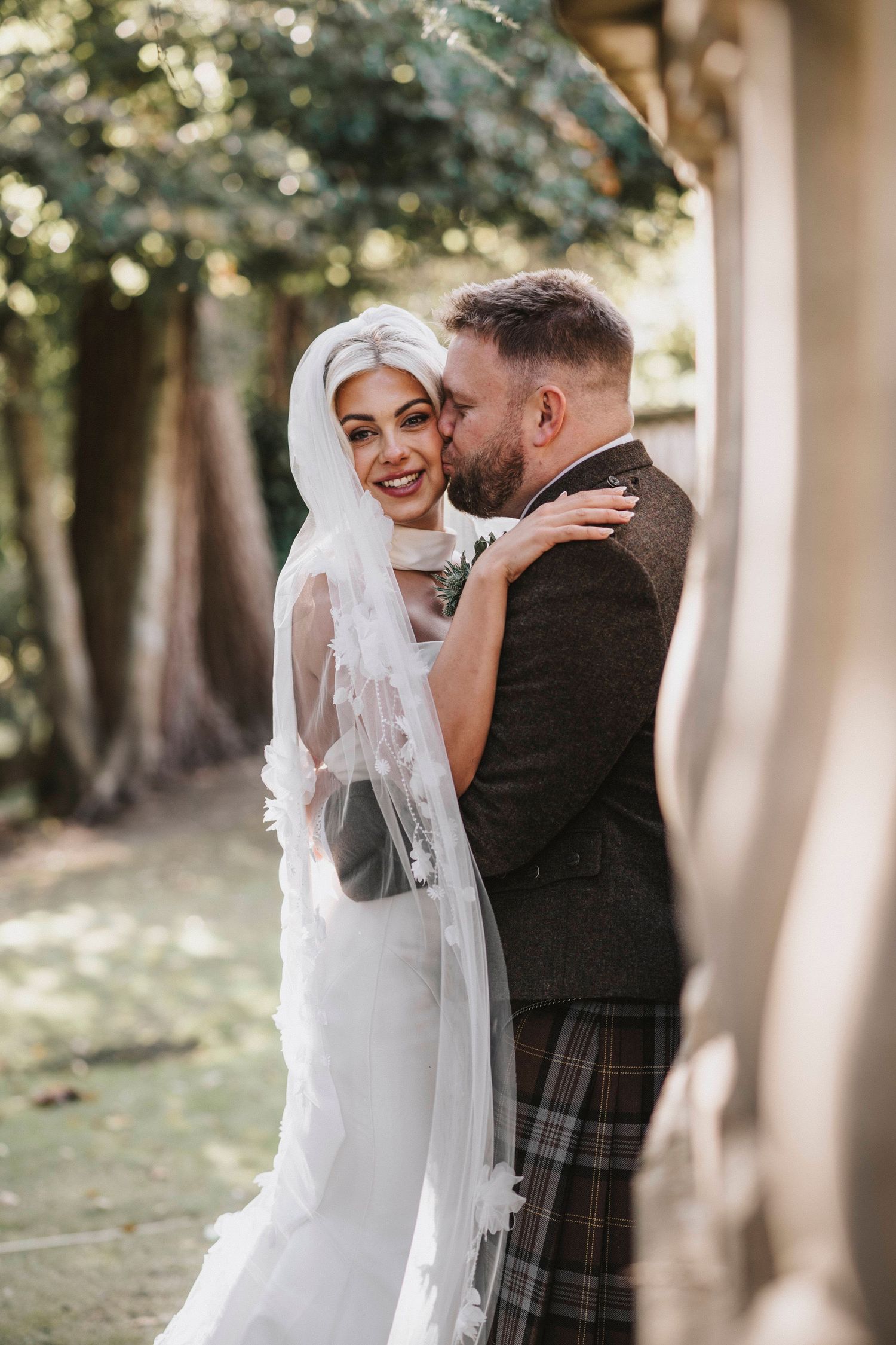 A romantic wedding couple embraces outdoors, with the groom wearing a traditional Scottish kilt and the bride in white.