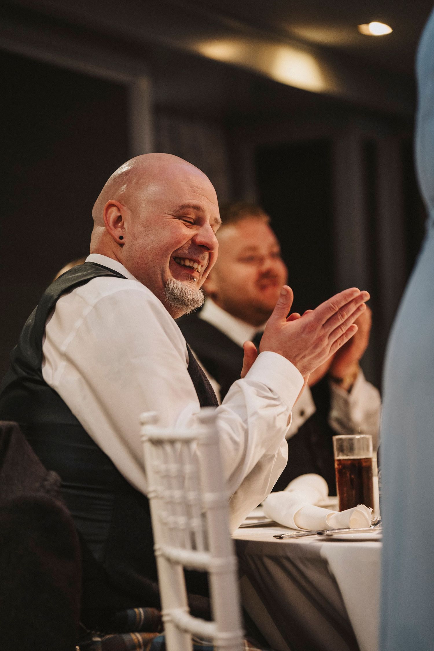 A guest at a wedding reception laughs heartily while clapping during dinner at an indoor venue.