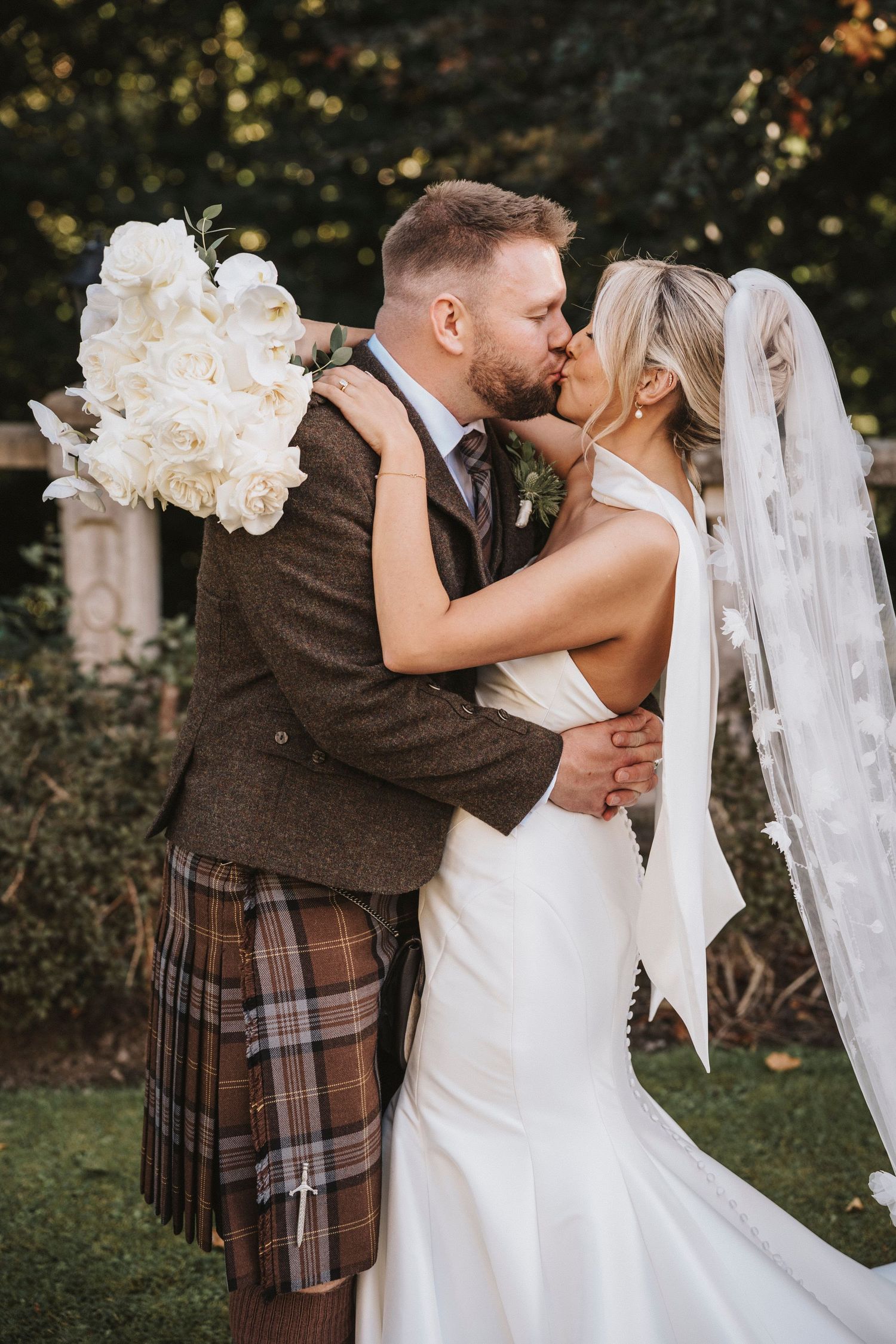A romantic wedding moment with a couple embracing, the groom wearing a traditional Scottish kilt and brown tweed jacket.