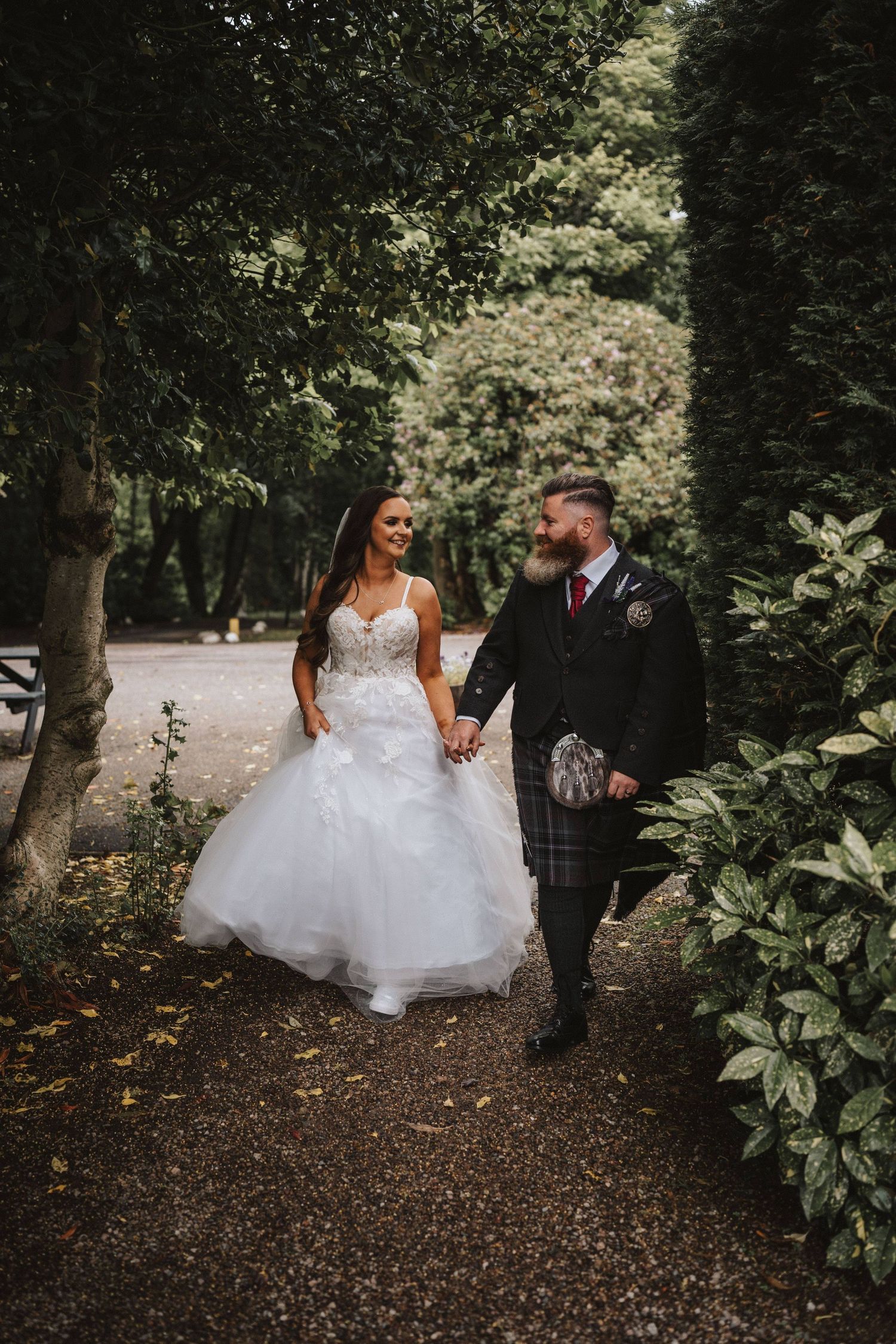 Couple walking through a wooded path surrounded by lush greenery and fallen leaves.