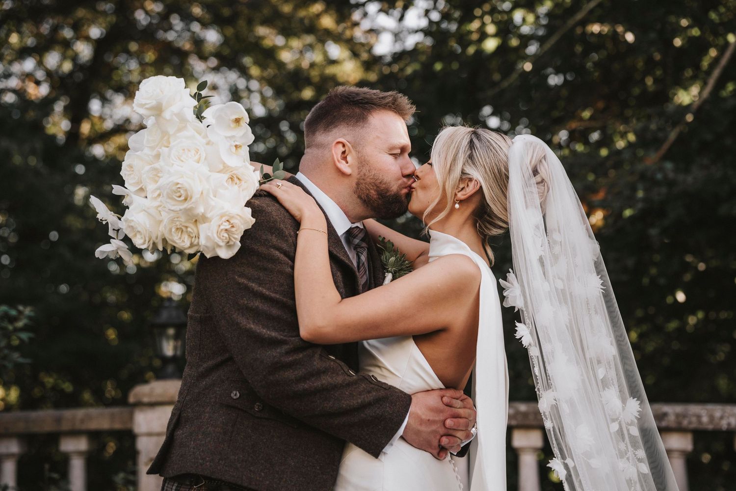 A romantic wedding couple embraces outdoors with a flowing white bouquet and veil against a natural backdrop.