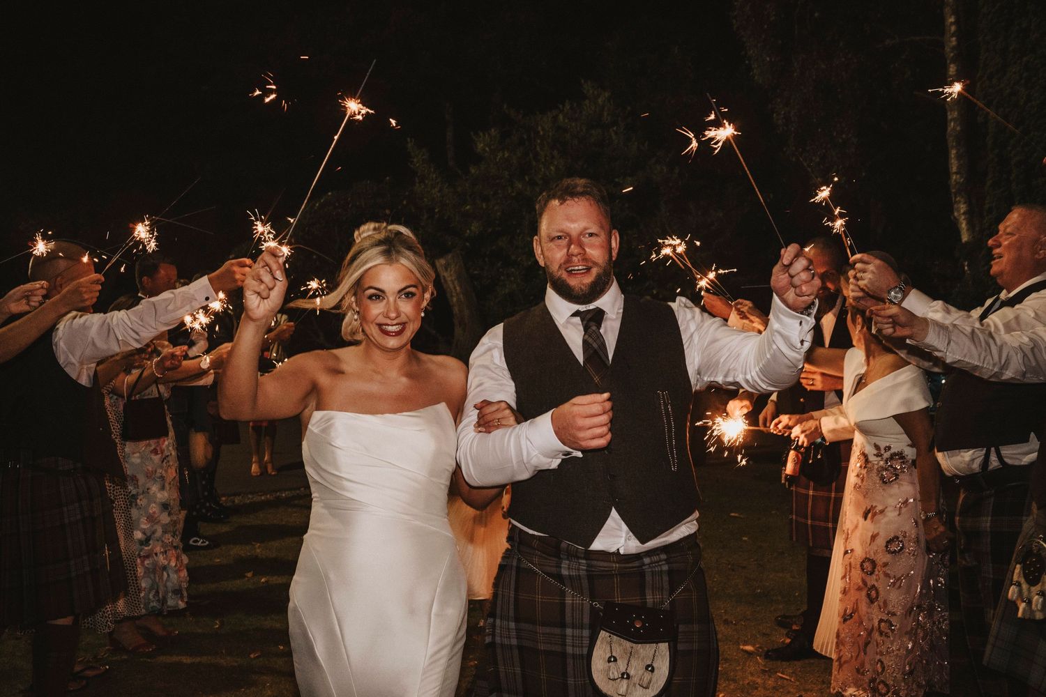 Wedding guests dance and celebrate with sparklers at an outdoor nighttime reception.