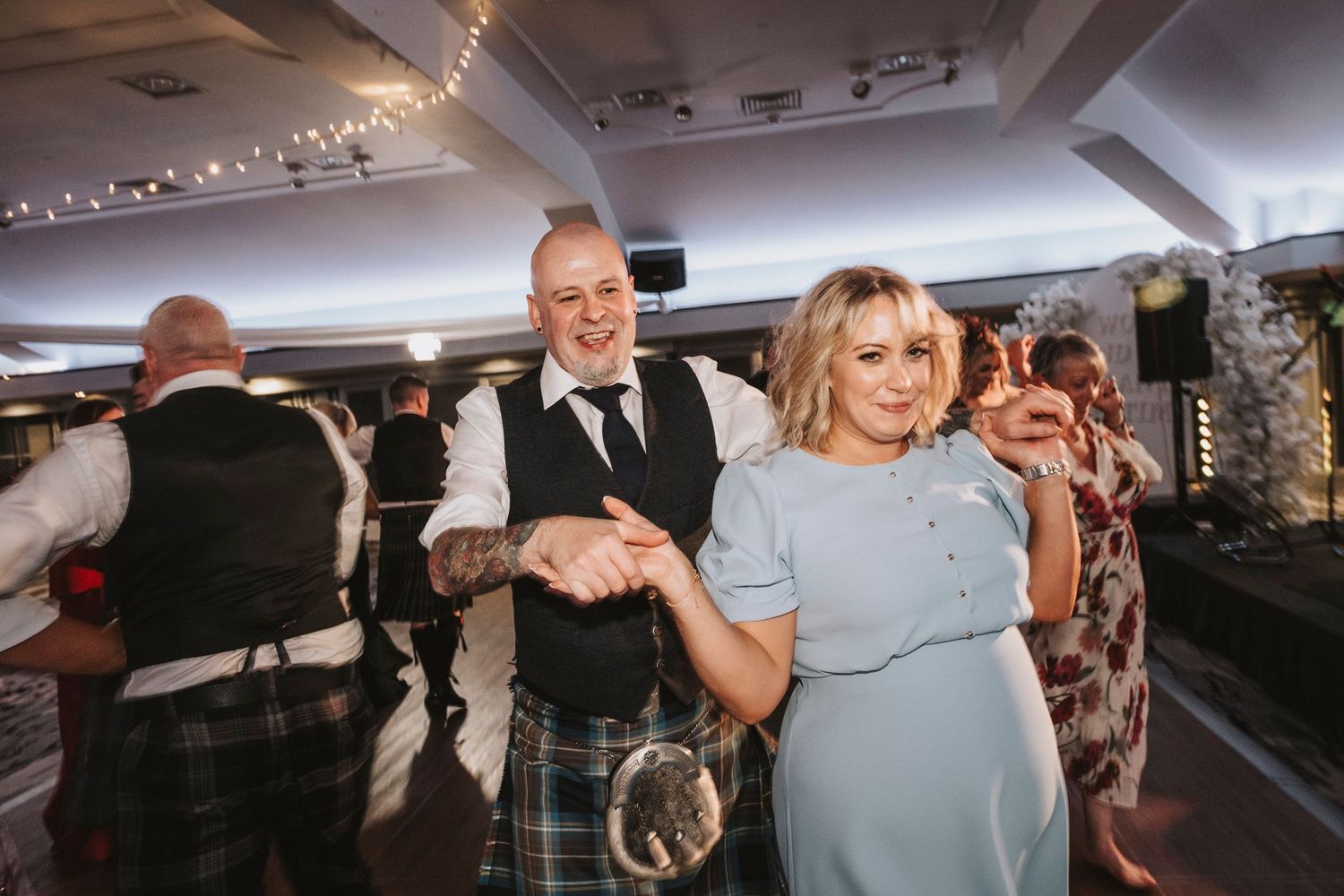 Wedding guests dancing and celebrating on a dance floor under string lights at an indoor reception venue.