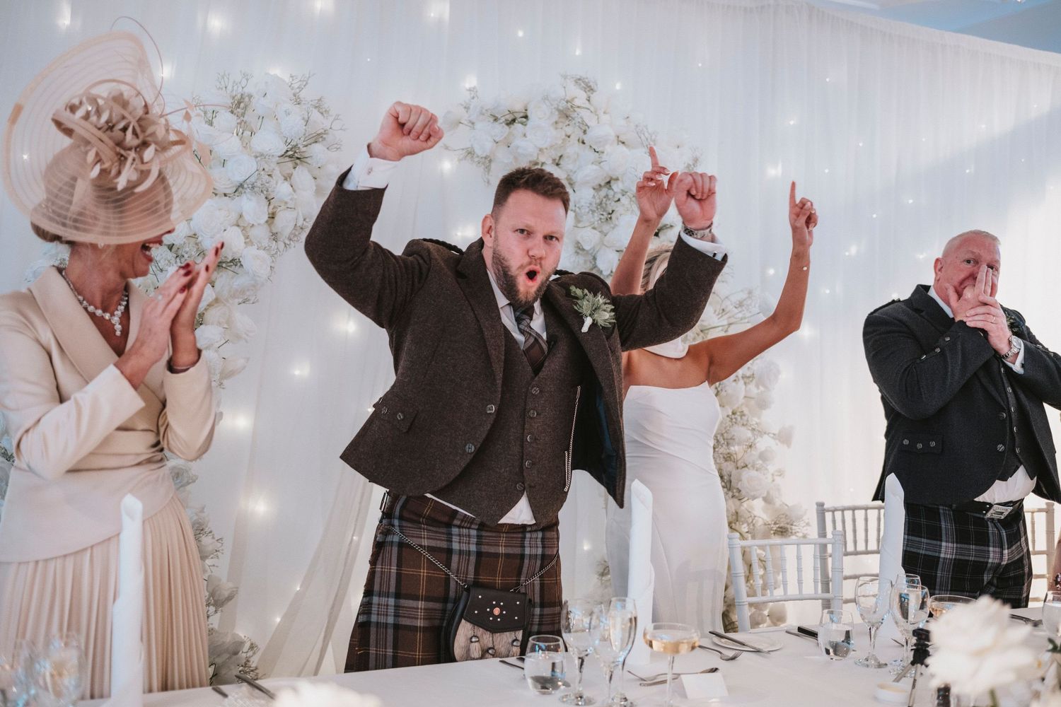 Wedding guests celebrate and dance enthusiastically at a reception with floral decorations and twinkling lights in background.