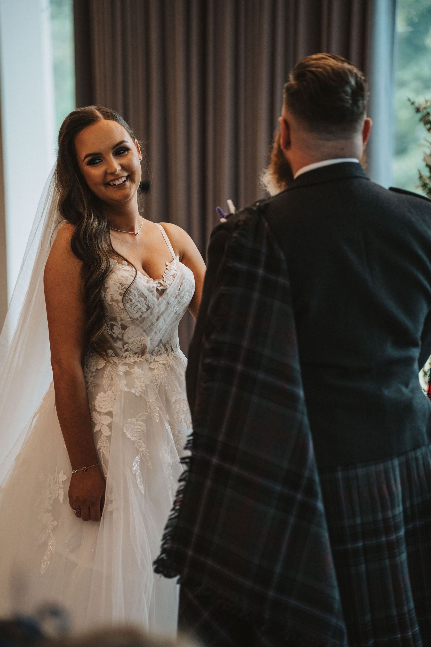 A bride in a white lace gown and groom in traditional Scottish kilt share candid moments during a wedding ceremony.