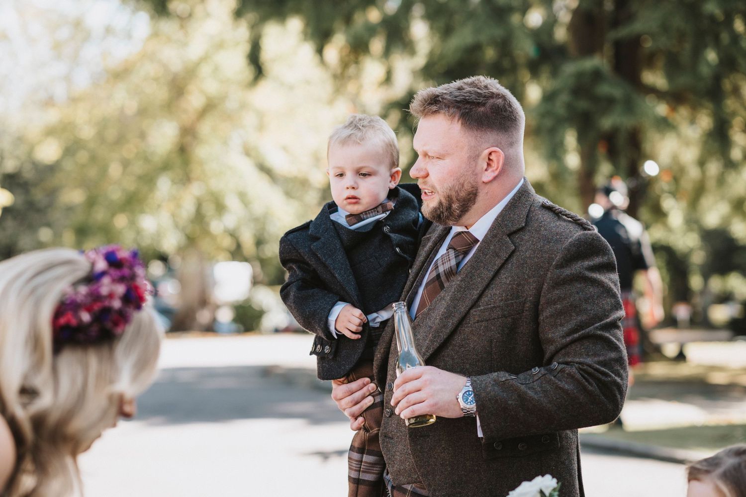 A well-dressed person in a tweed suit holds a small child outdoors on a sunny day in a park setting.