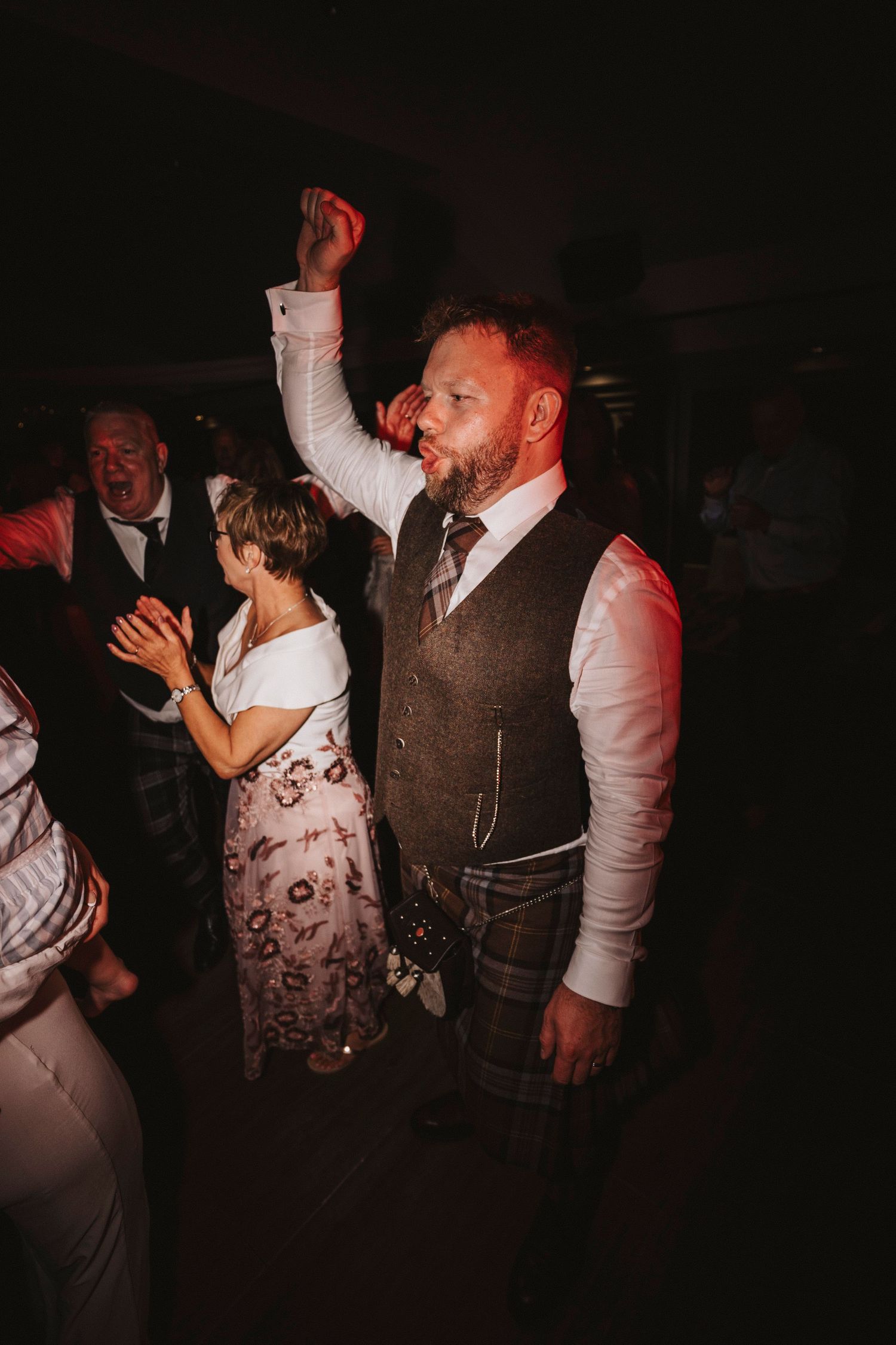 Wedding reception guests dance and celebrate in a dimly lit venue with red lighting accents.