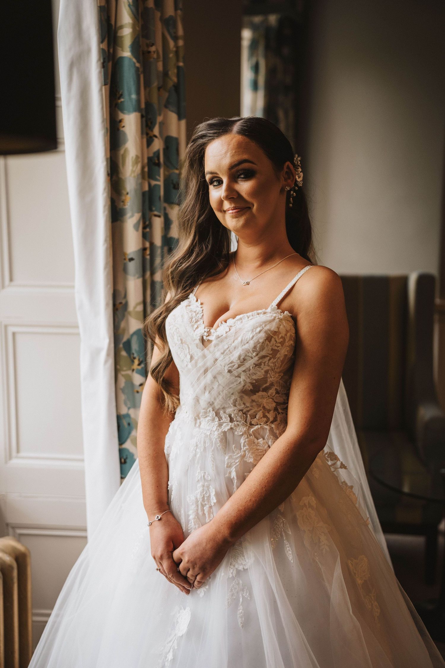 A close-up view of a bride in a white lace wedding gown with delicate details and embellishments against a dark room.