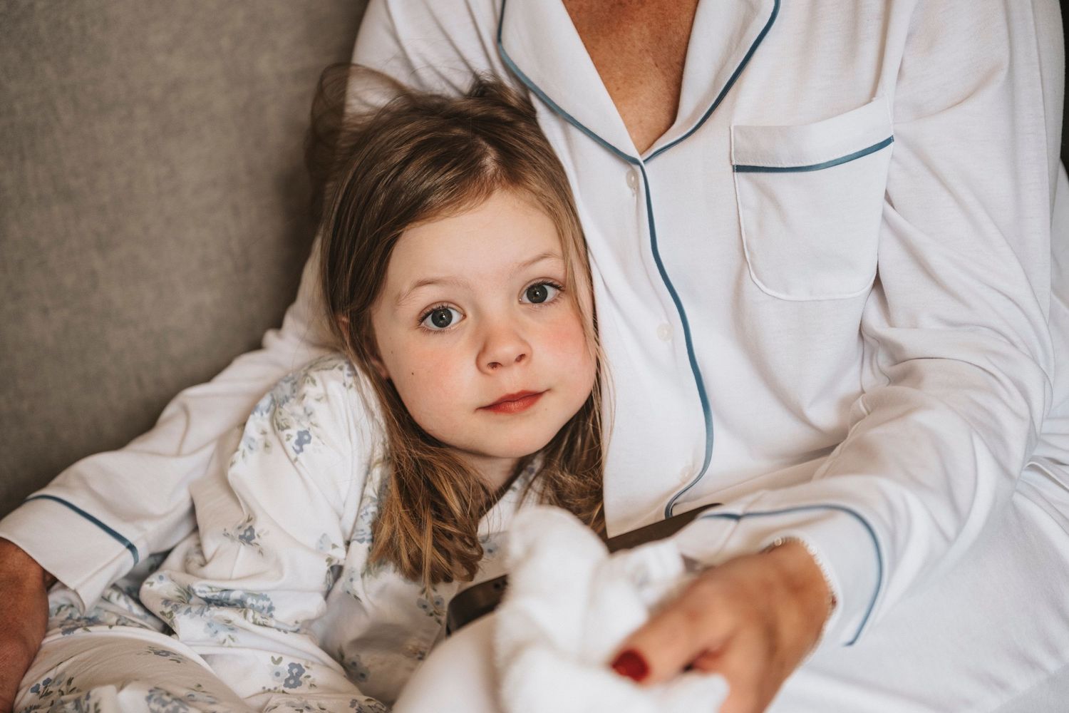 A young child in white pajamas cuddles close to someone wearing piped pajamas while relaxing on a couch.