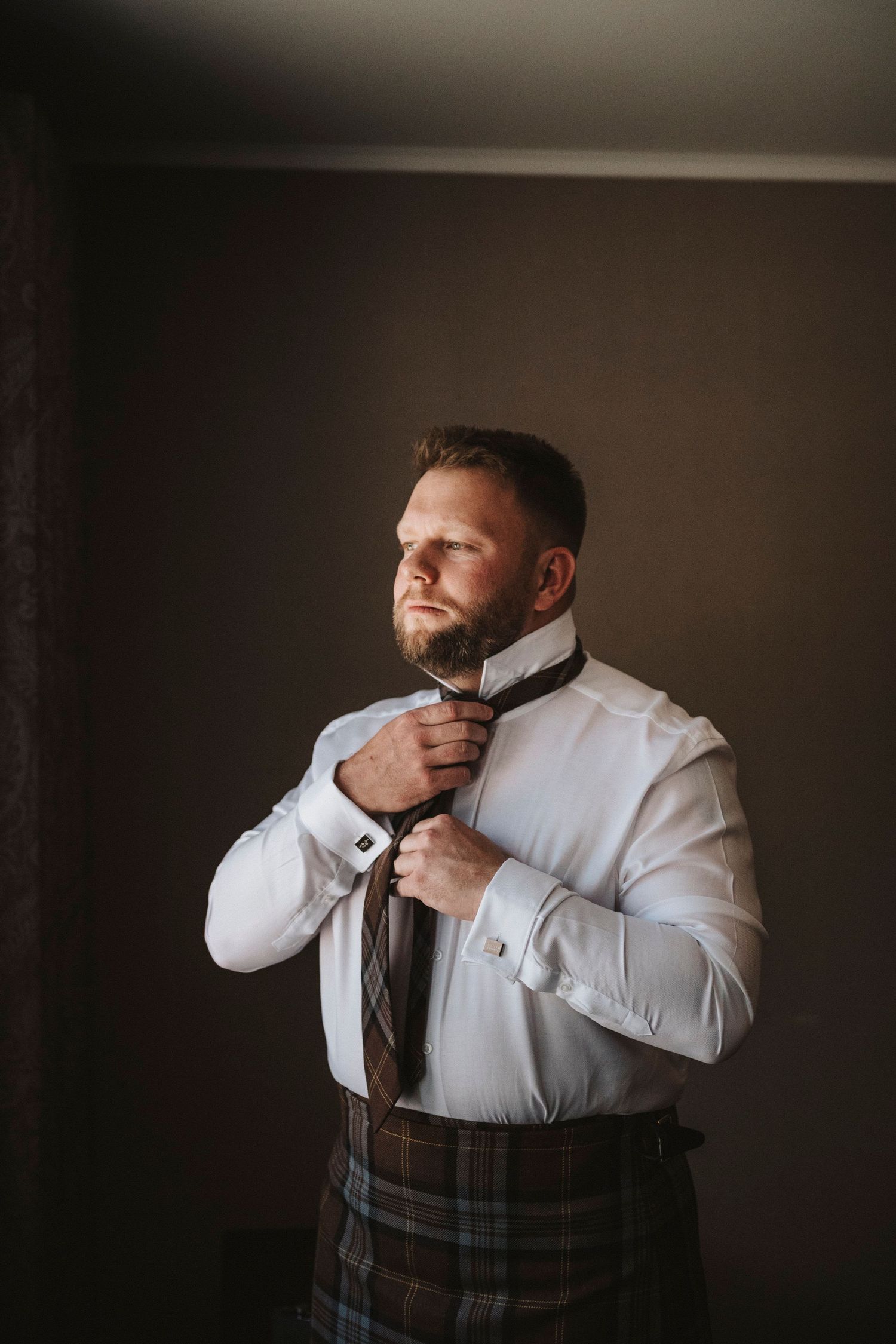 A person in traditional Scottish attire adjusts their tie while wearing a crisp white dress shirt and plaid kilt.