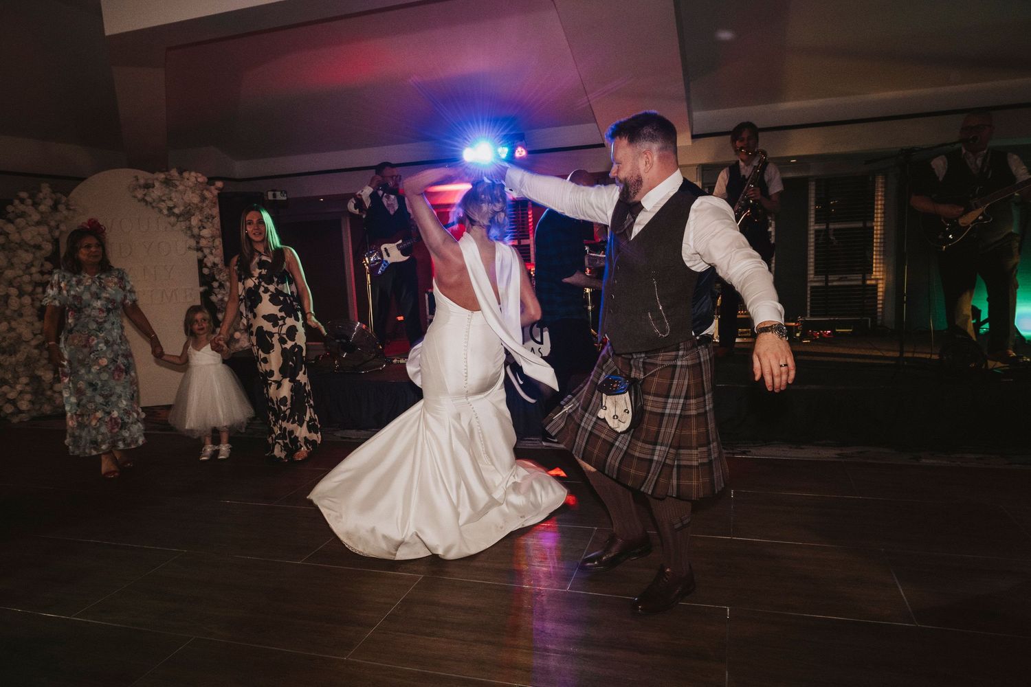 Wedding guests dance and celebrate on a dimly lit dance floor with colorful lights in the background.