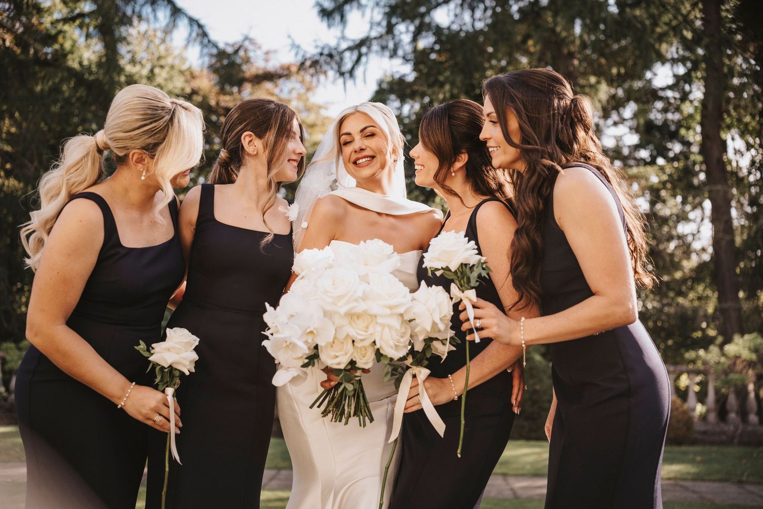 Bridal party in black dresses shares a joyful moment with bride in white holding large white bouquet outdoors.