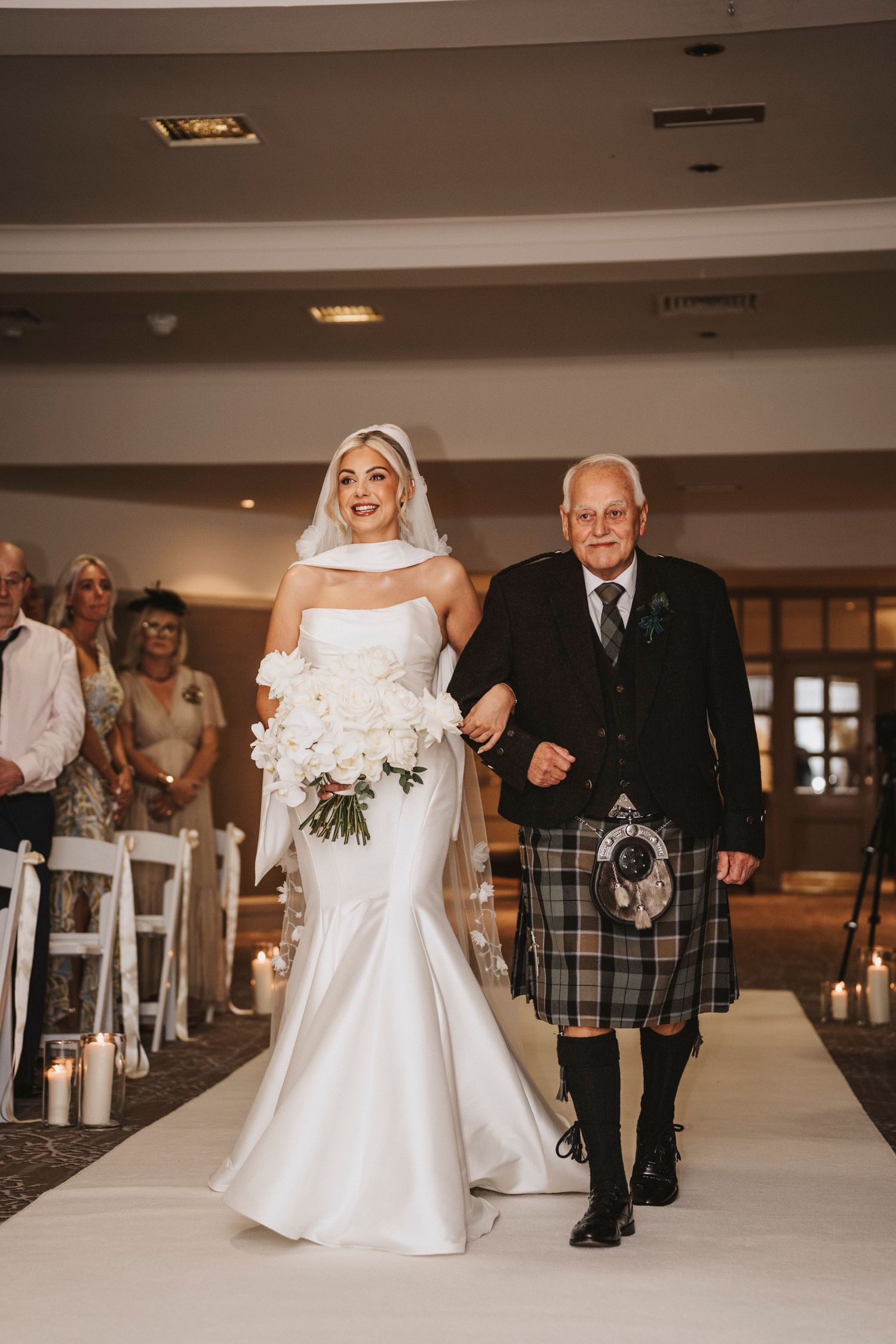 A bride in a white mermaid gown walks down the aisle with an older gentleman wearing a traditional Scottish kilt.