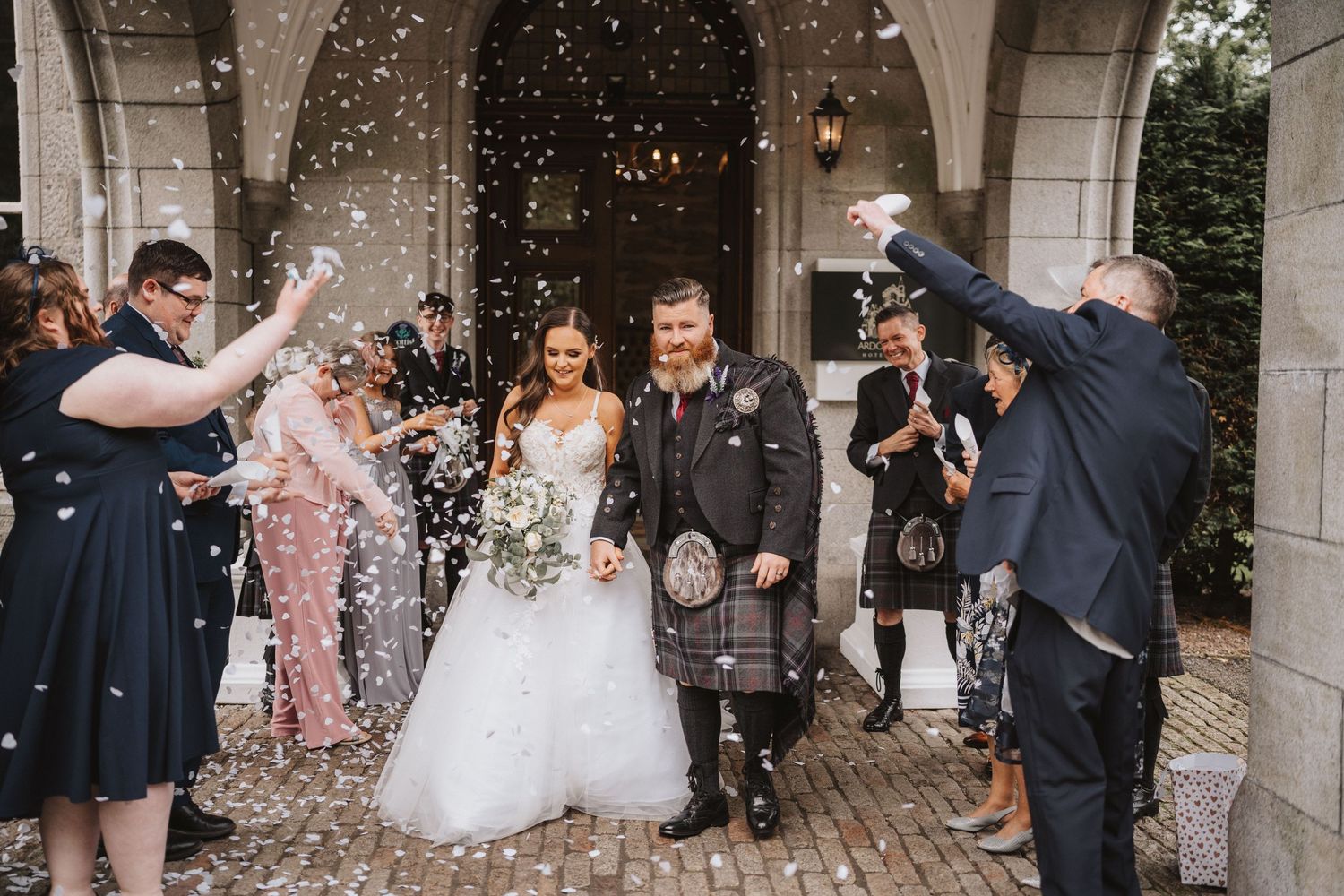 Wedding celebration with guests showering newlyweds in confetti while exiting stone archway.