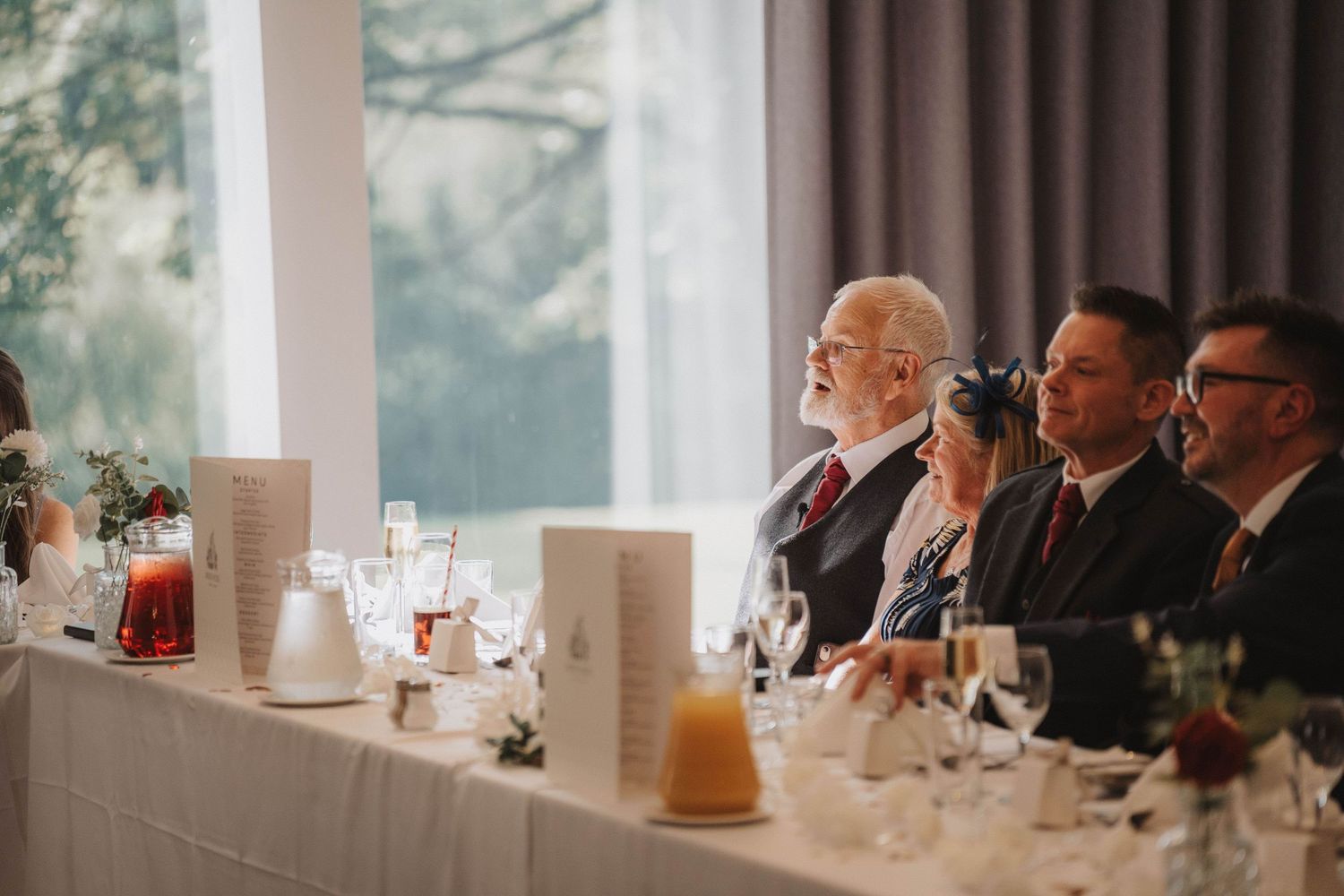 Long dining tables with white linens and centerpieces set up beside large windows in an elegant reception hall.