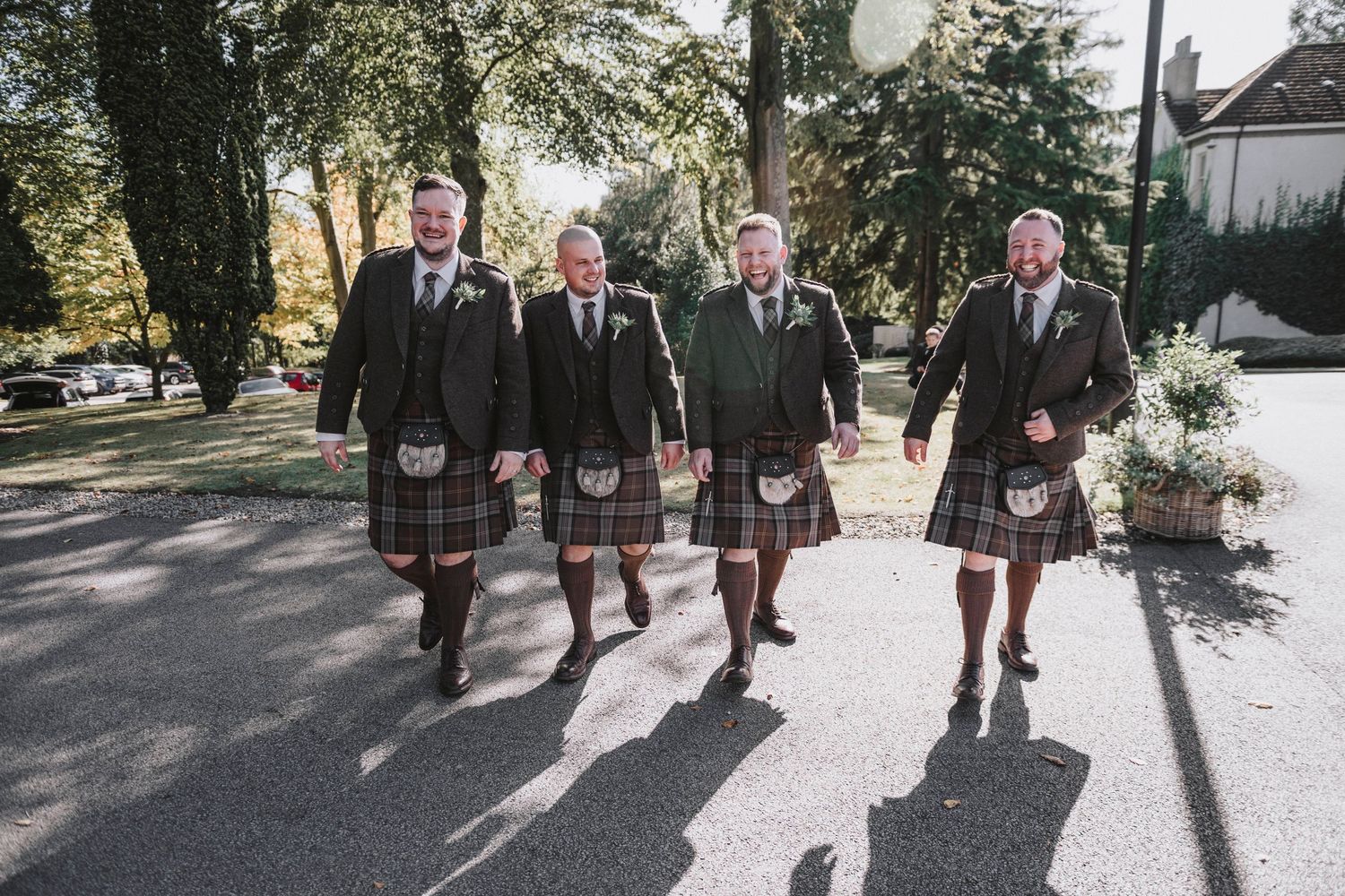 A wedding party in traditional Scottish kilts walking together on a sunny day through a tree-lined path.