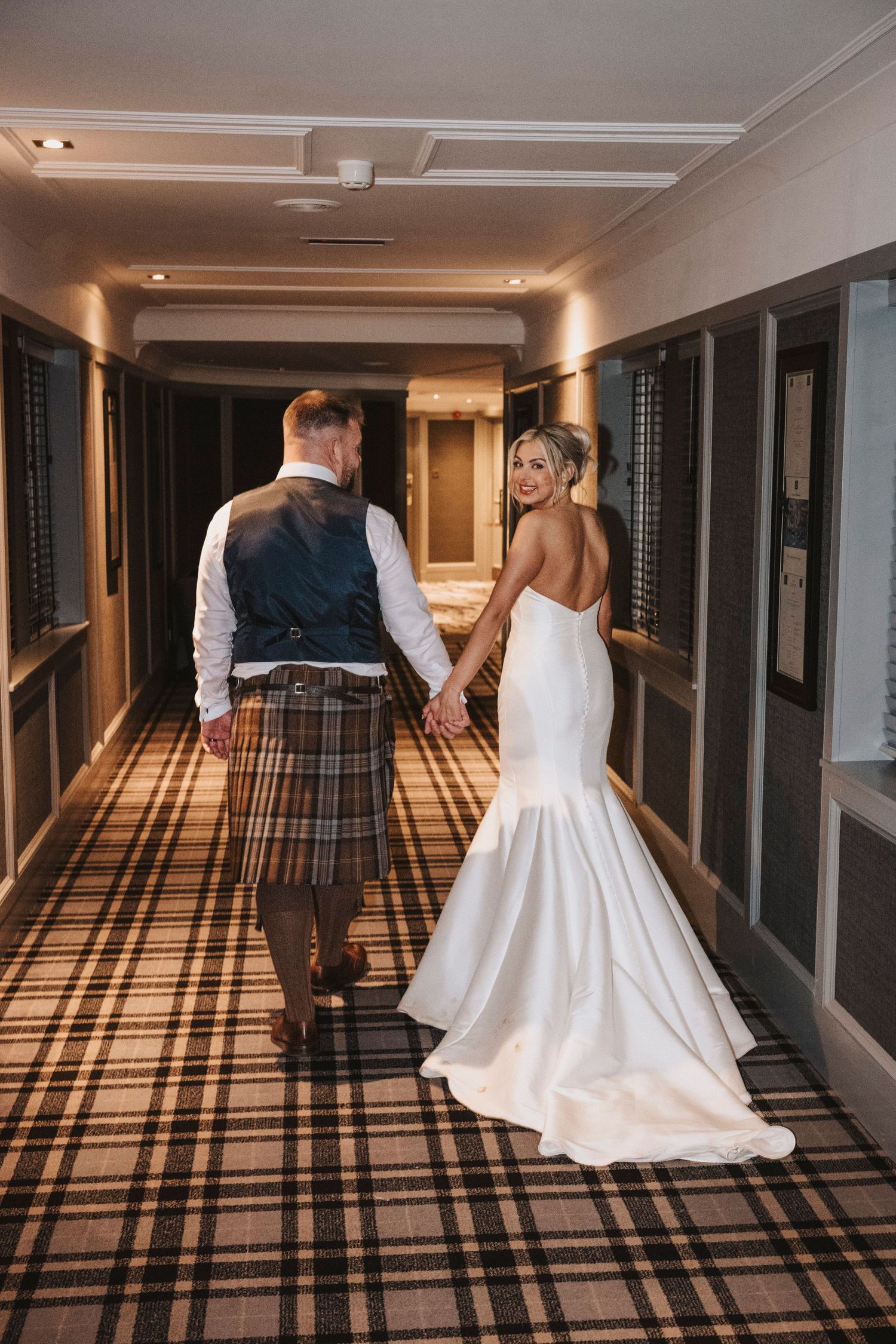 A couple in wedding attire walks down a carpeted hallway with dramatic lighting casting geometric shadows.