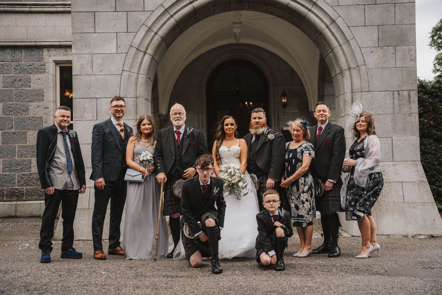 Wedding party group poses in front of gray stone archway outdoors.