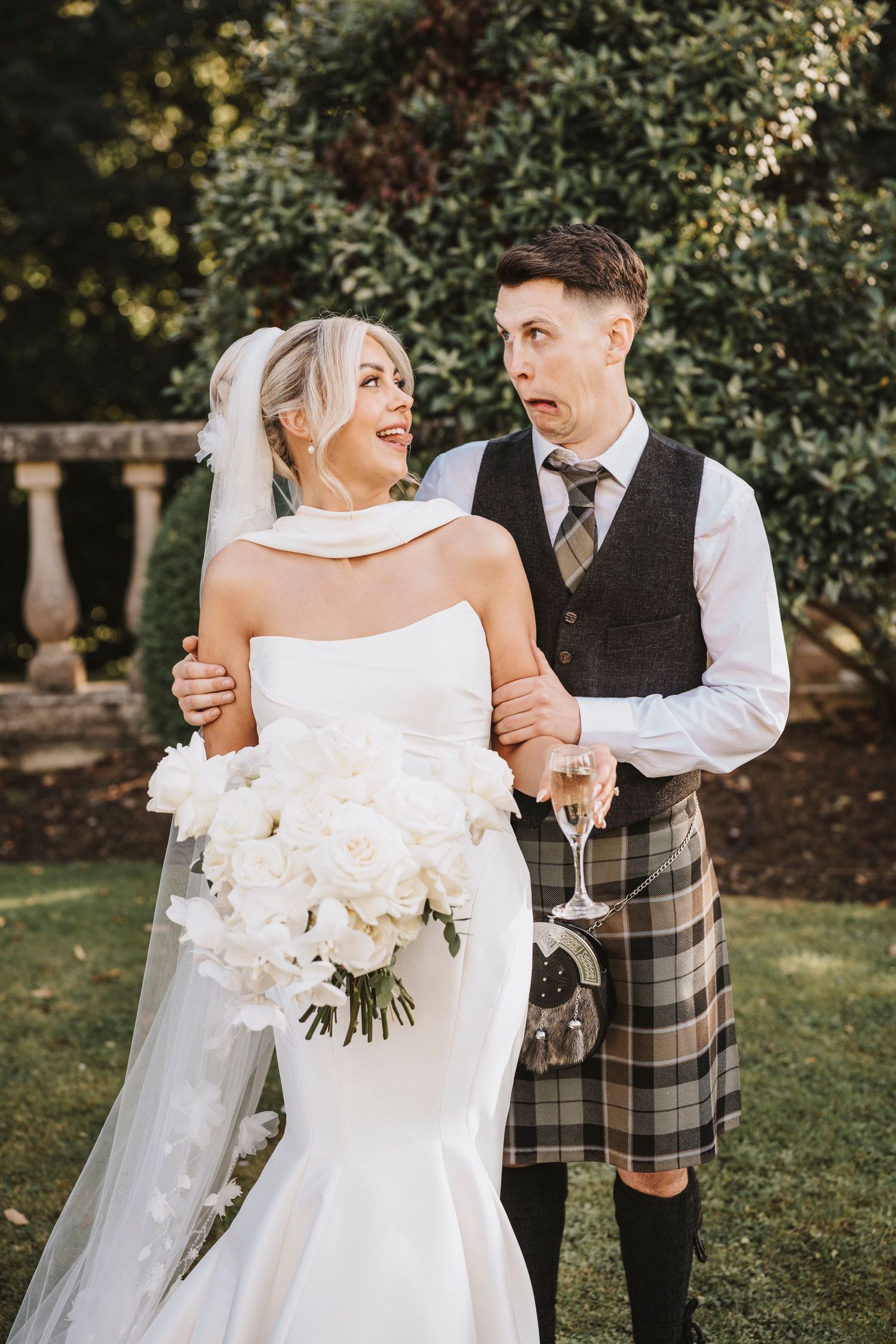 A romantic wedding couple embraces outdoors with the bride in white and groom wearing a traditional Scottish kilt.