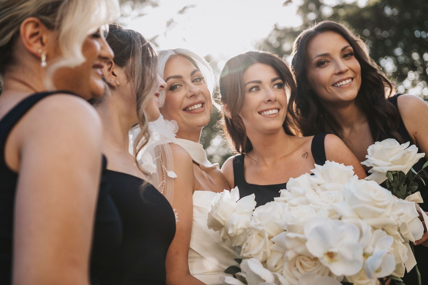 Wedding party members smile and laugh together while holding white floral bouquets during an outdoor celebration.