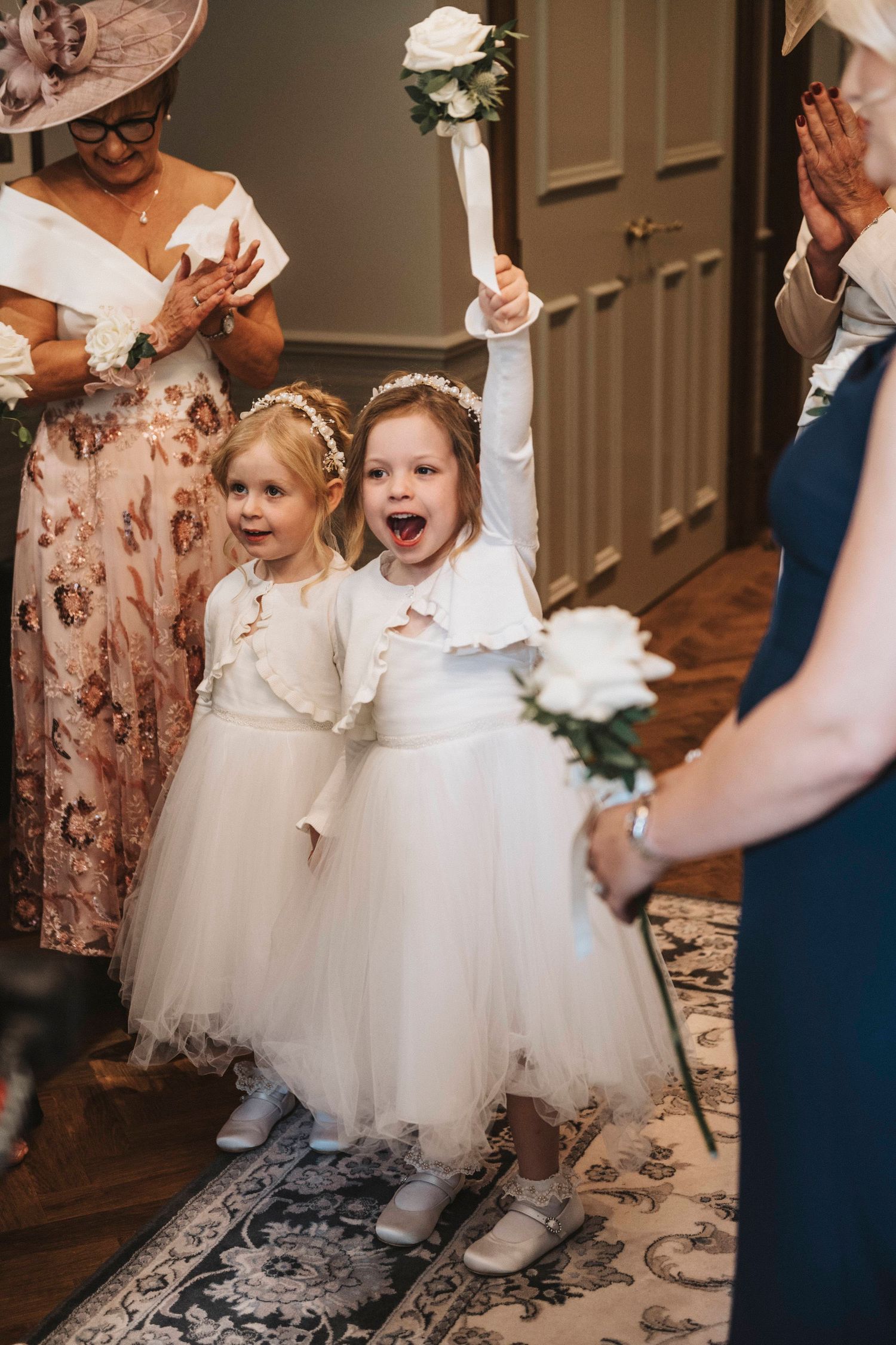 Two flower girls in white tulle dresses dance excitedly on a dark floor during a wedding celebration.