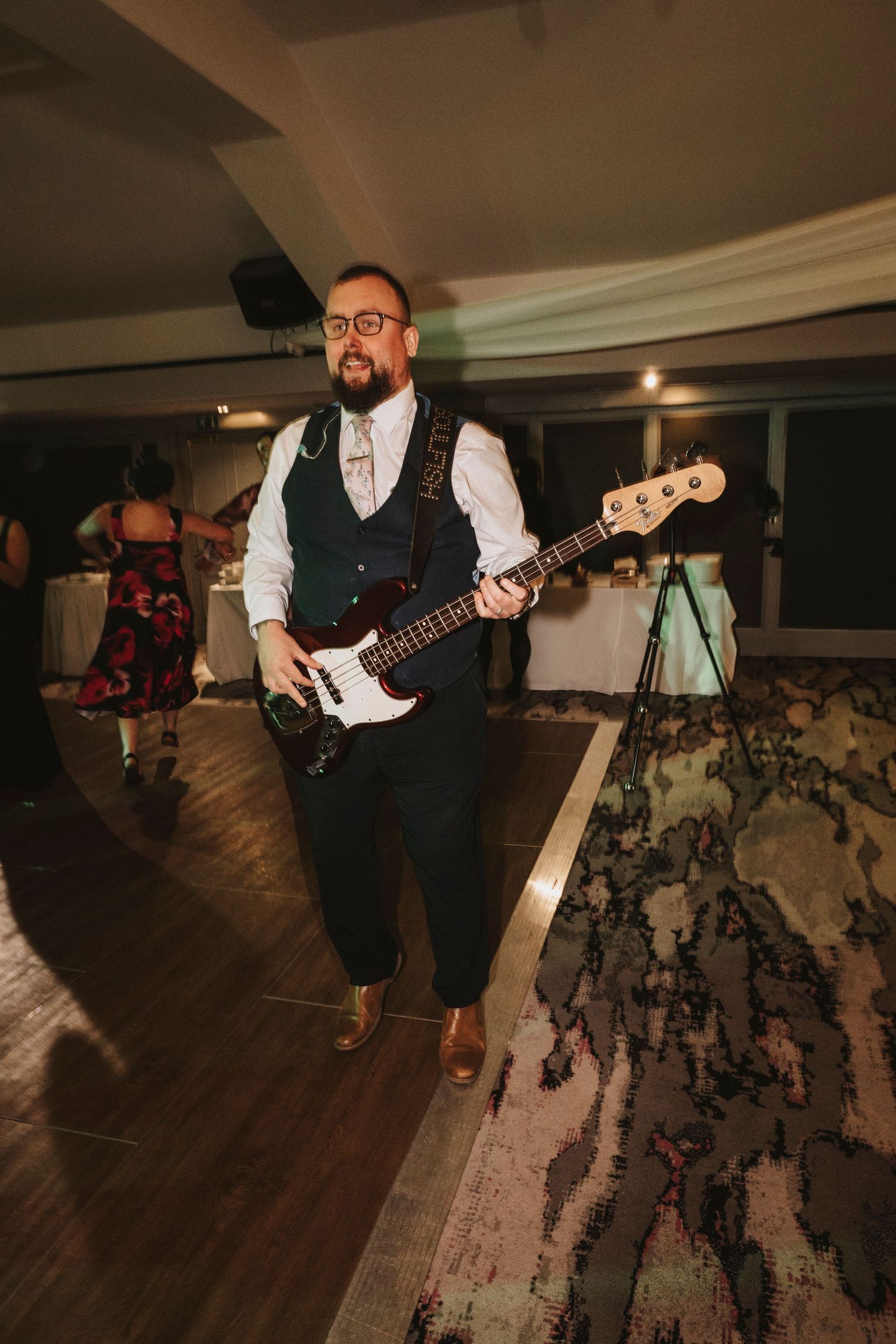 A musician in a vest plays electric bass guitar on a wooden dance floor during a performance.
