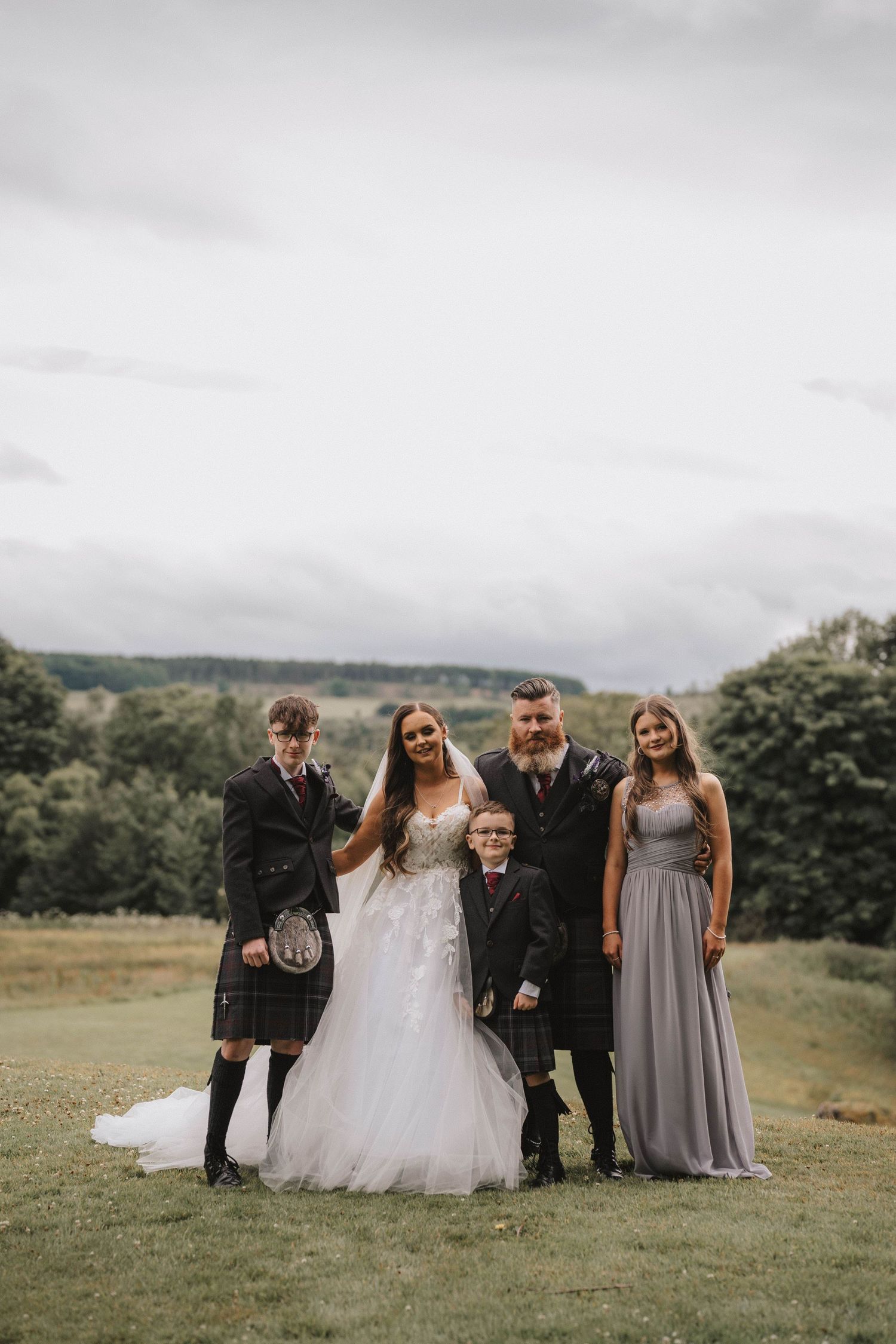 Wedding party poses for group photos on green hillside with overcast sky in background.