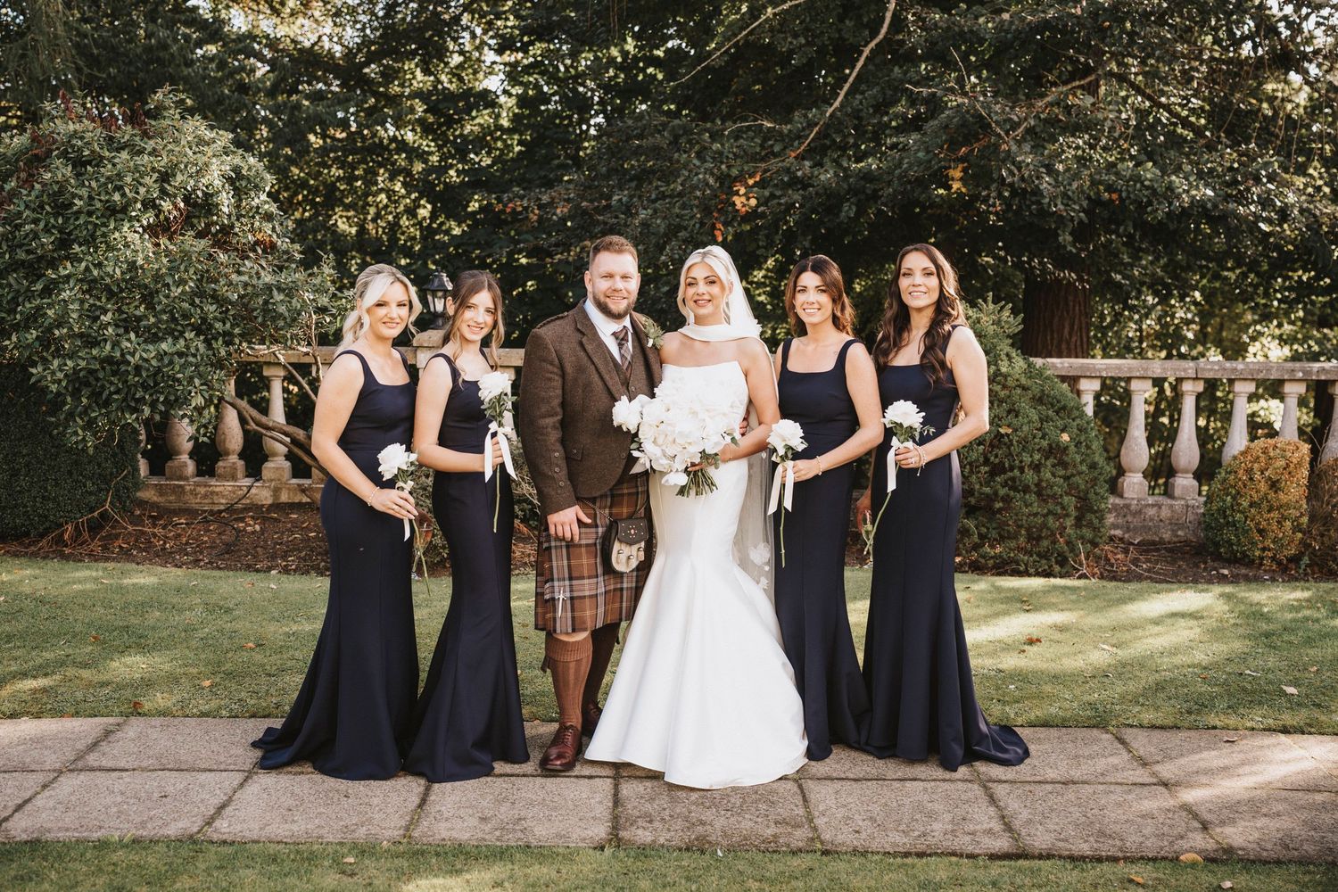 A wedding party poses outdoors in formal attire with bridesmaids in navy dresses against a stone balustrade and garden backdrop.