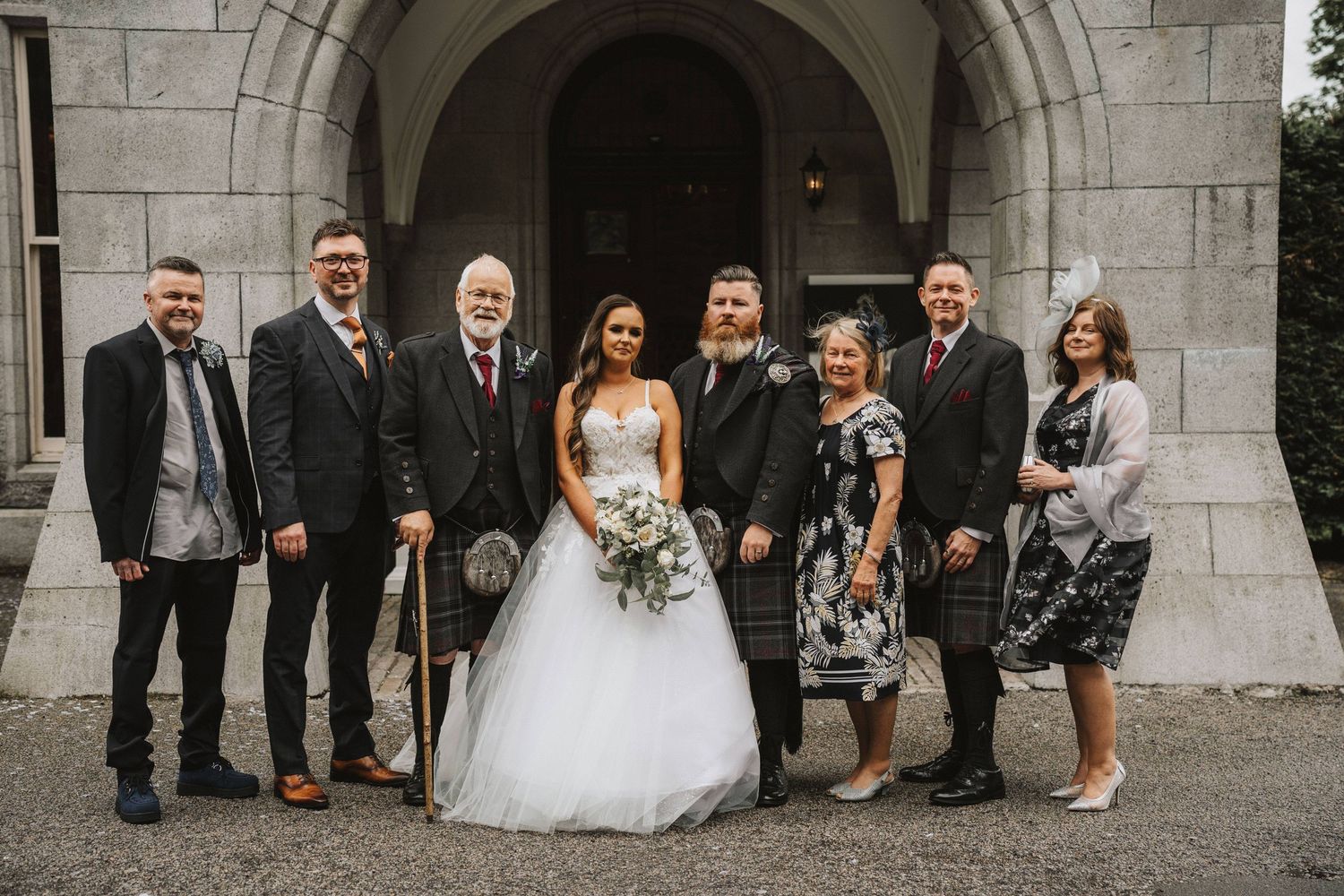 Wedding party members pose together in front of a stone archway during an outdoor celebration.