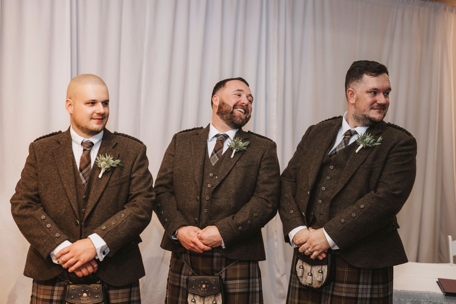Groomsmen in brown suits and ties stand in a row at a formal wedding ceremony.