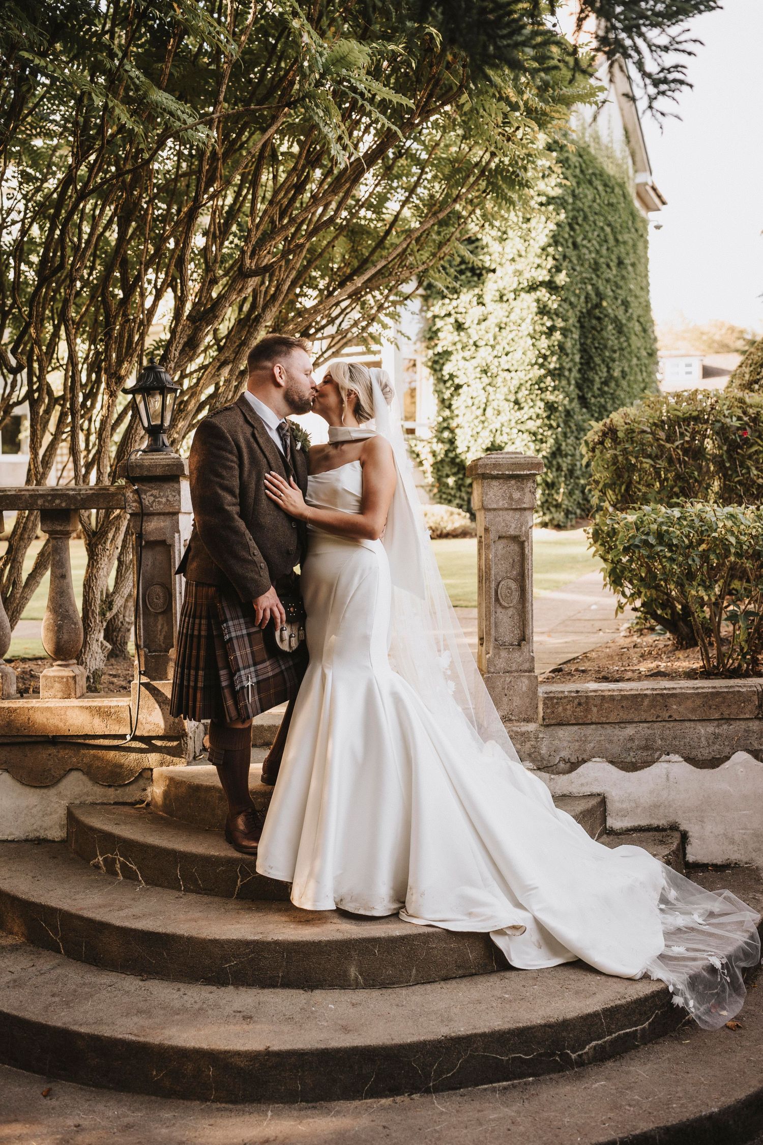 A couple in wedding attire share intimate moments on curved stone steps surrounded by lush greenery at an outdoor venue.