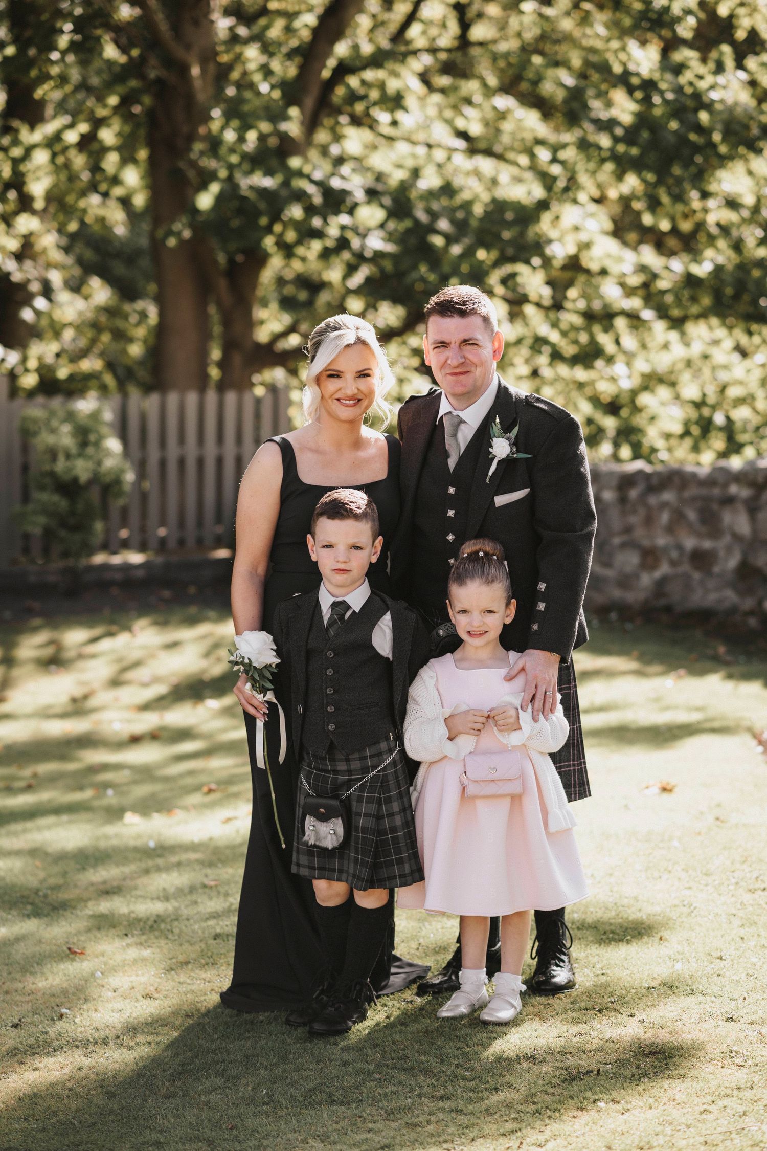 A family in formal Scottish attire poses together on a sunny lawn during a special occasion.
